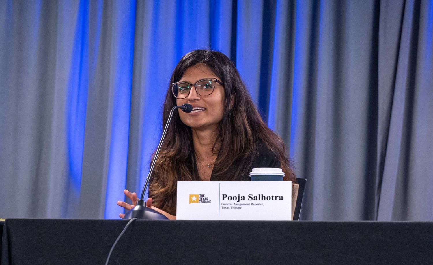 A speaker stands at a podium during a presentation with blue curtains in the background.