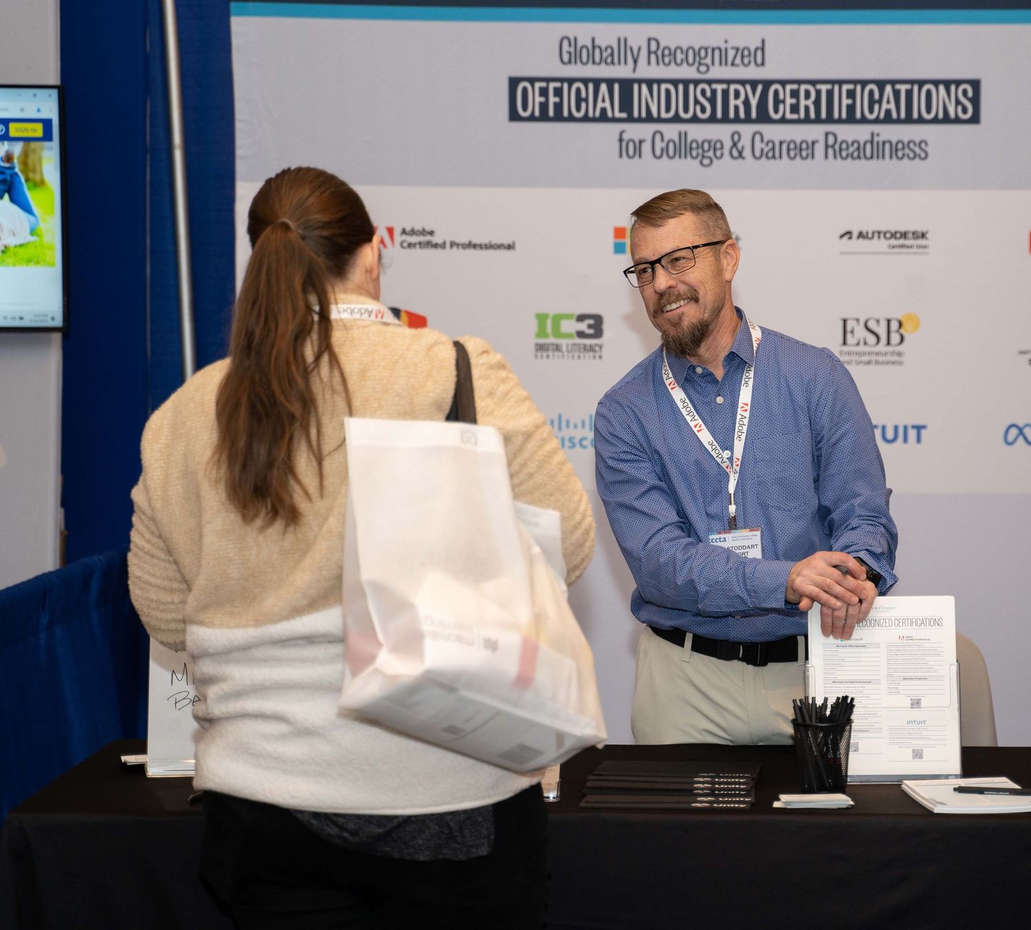 Business professionals signing documents at a certification event with banners and displays visible in the background.