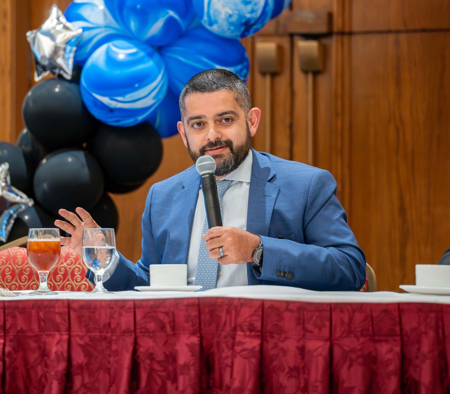 Speaker in a blue suit jacket holds a microphone while seated at a table with red tablecloth and blue balloons in background.