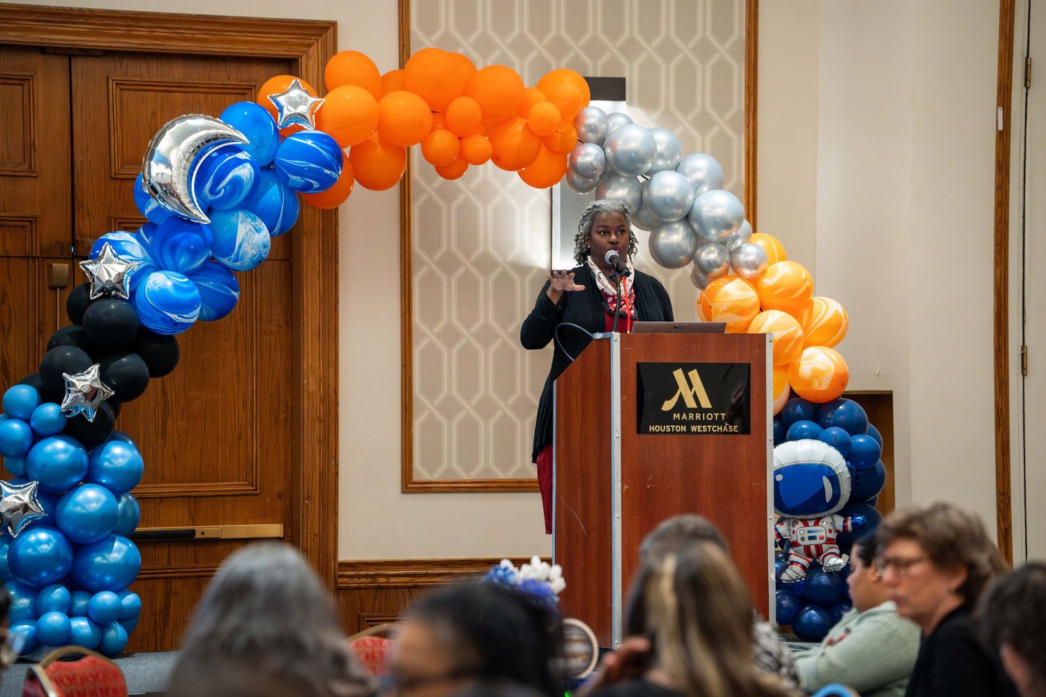 Attendees gather in a banquet hall decorated with festive blue and orange balloon arrangements for a celebration.