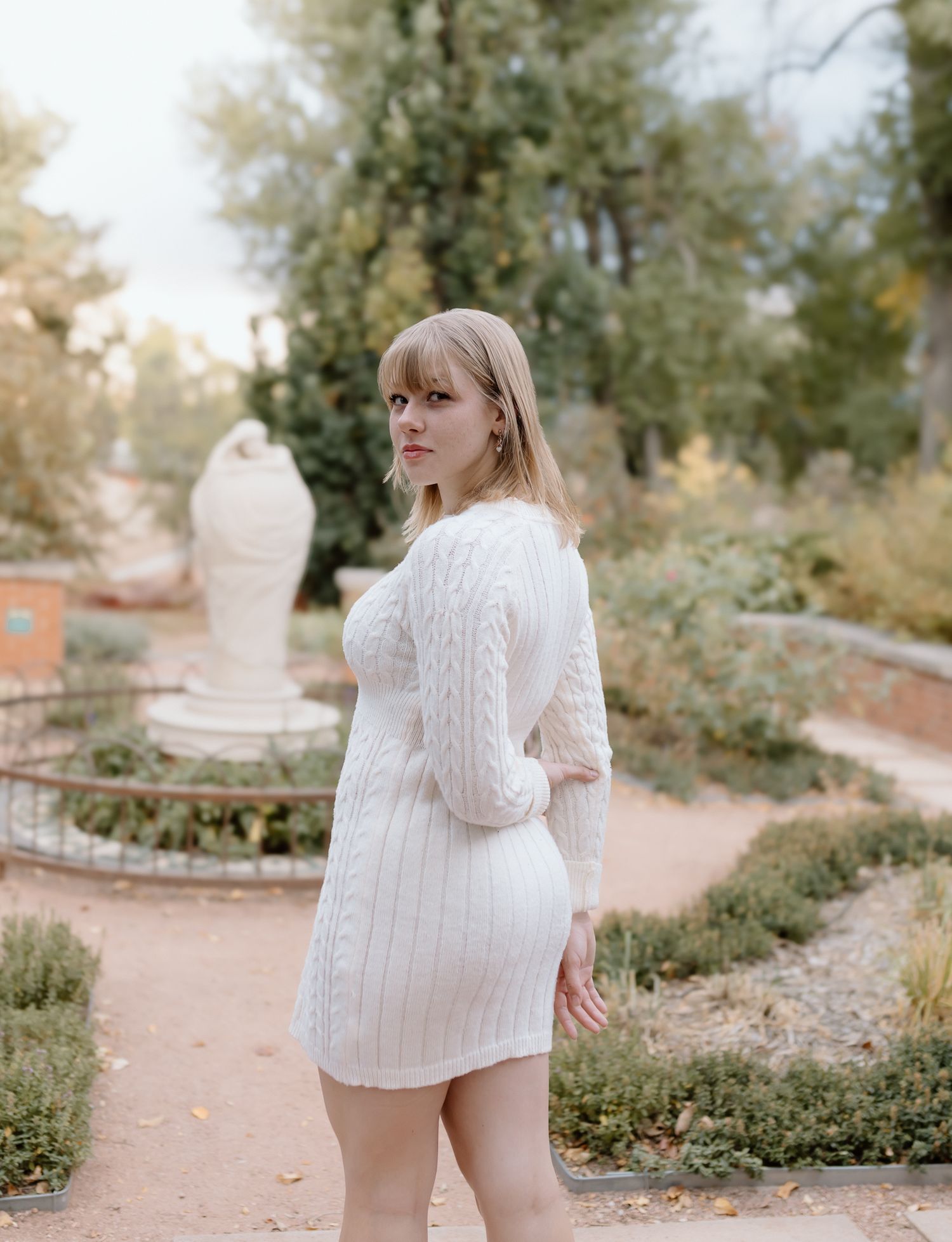 A person in a cozy white cable knit sweater poses in a garden setting during autumn.