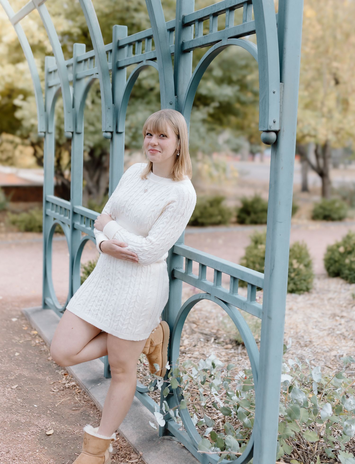 A person in a white sweater and shorts poses playfully on a blue metal railing structure in an outdoor setting.
