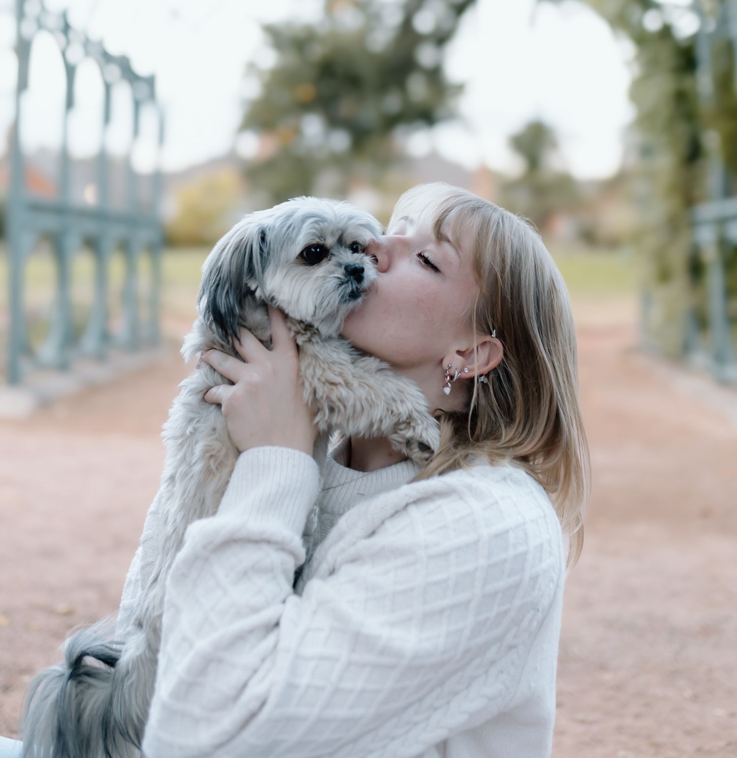 Small white dog being lifted and kissed by someone wearing a cream sweater in an outdoor setting.