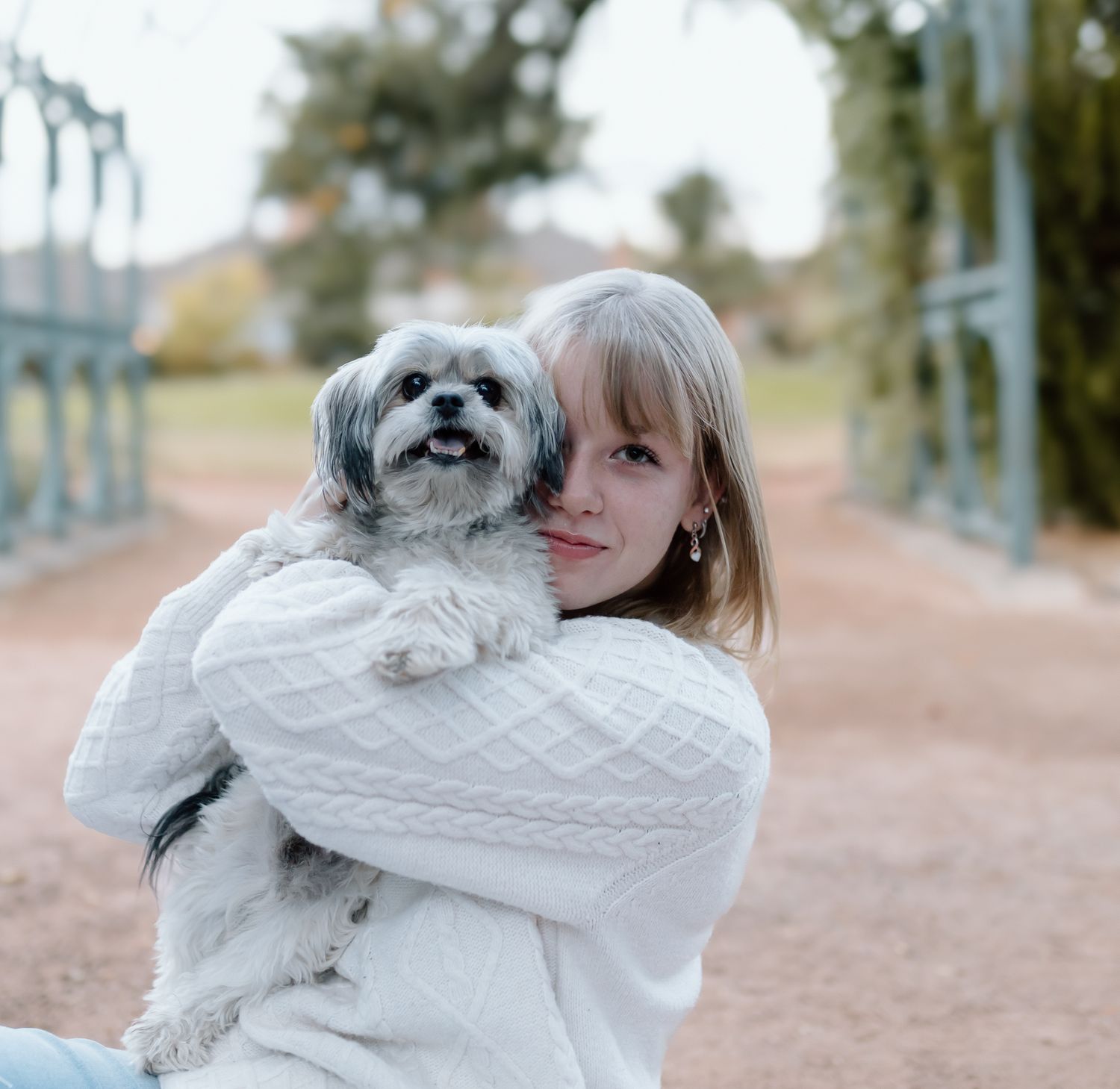 A person in a white sweater holds and cuddles with a small fluffy gray dog on a dirt path outdoors.