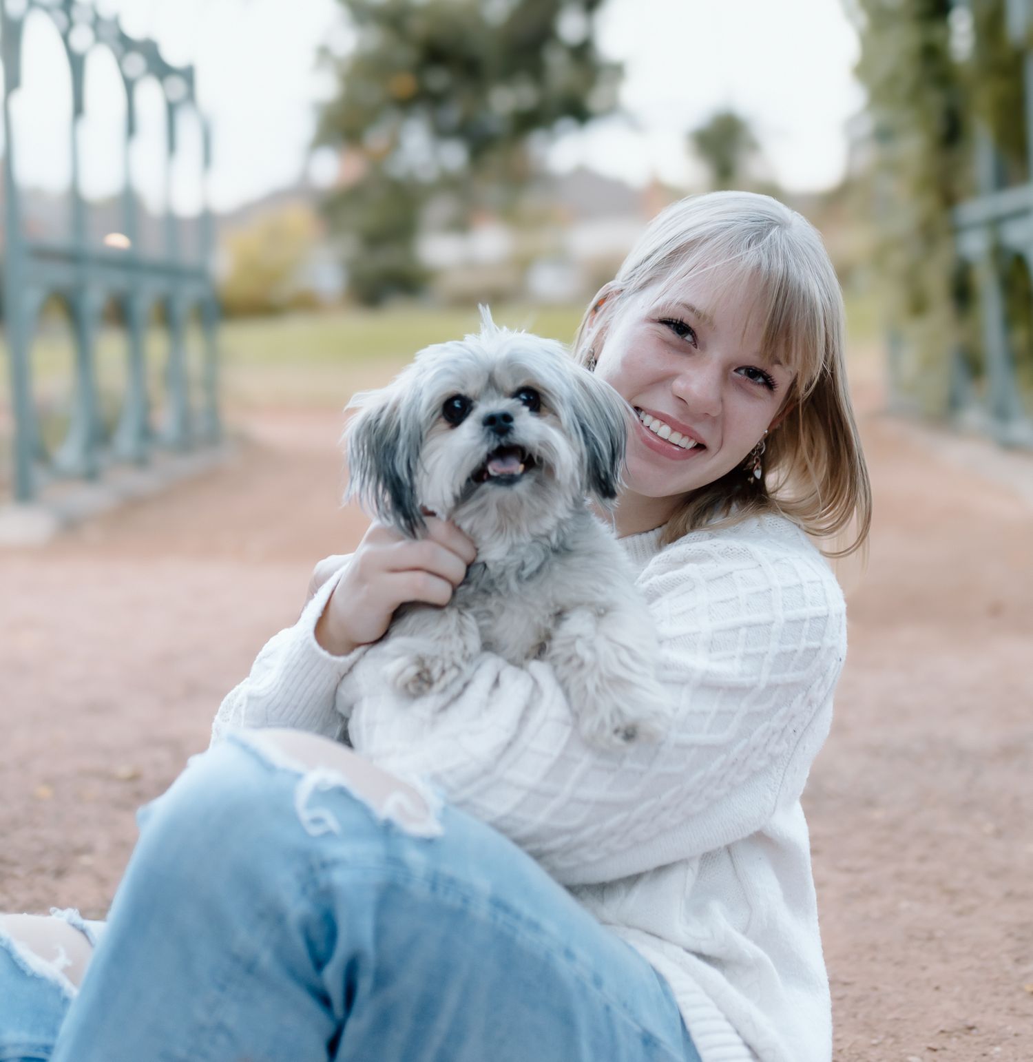 A person in a white sweater cuddles with a small fluffy white dog outdoors on a path.