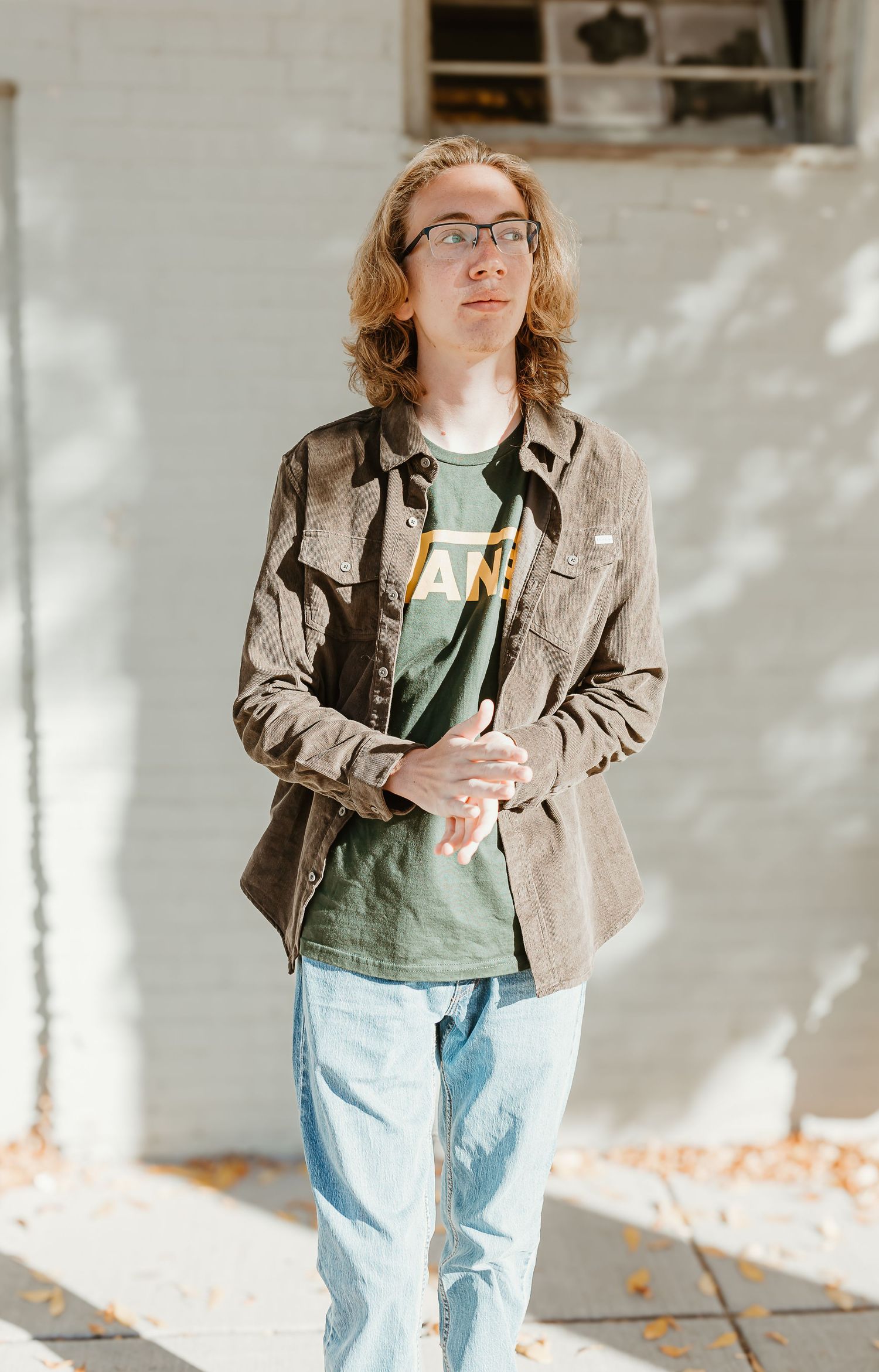 A person in a brown jacket and jeans poses casually against a sunlit wall in an indoor setting.