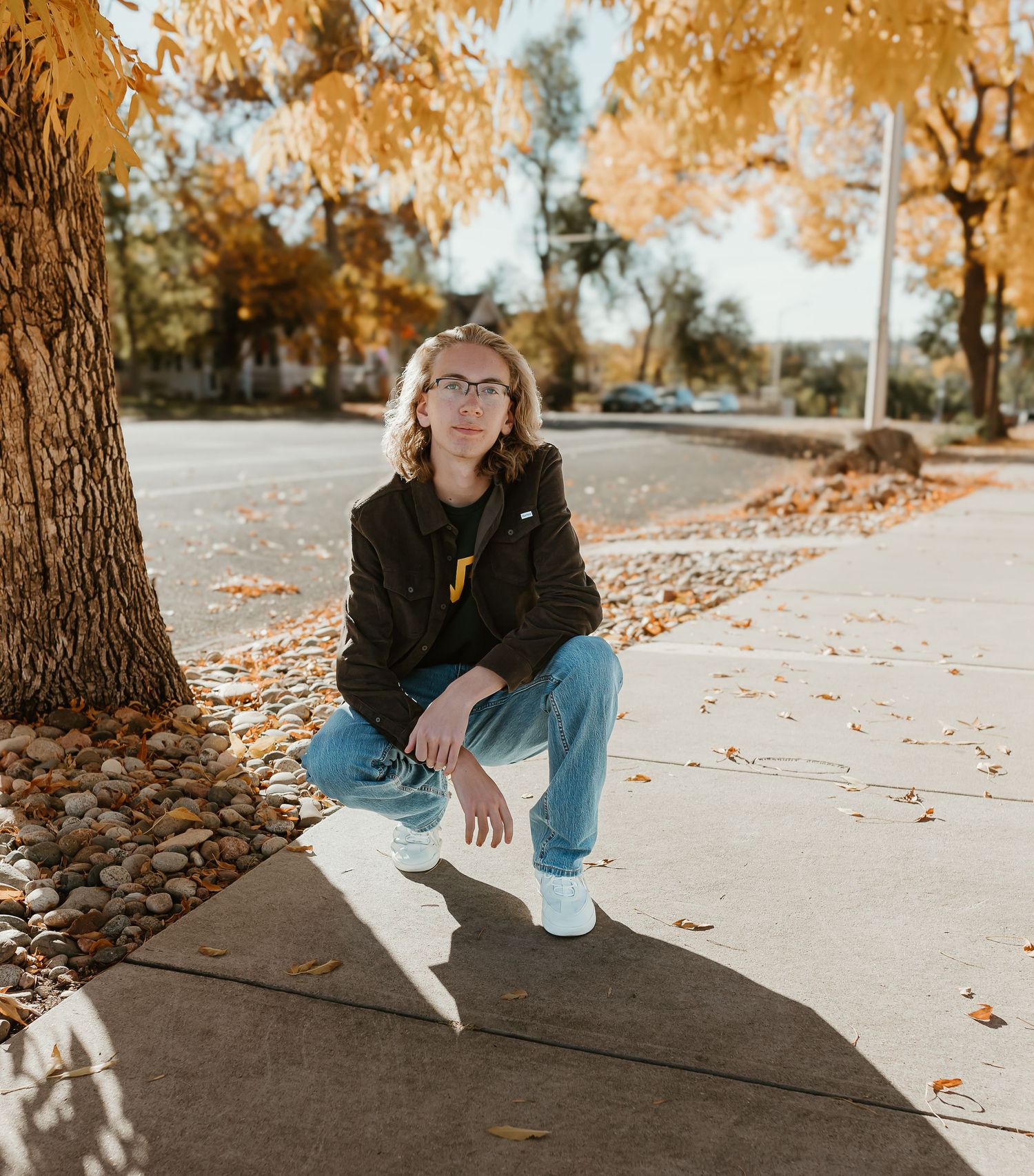 Person in casual attire sits on ground surrounded by autumn leaves and trees with warm golden lighting.