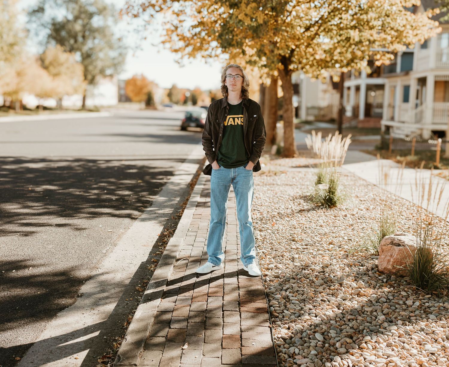A person in blue jeans and green sweater stands on a sidewalk surrounded by golden autumn leaves and dappled sunlight.
