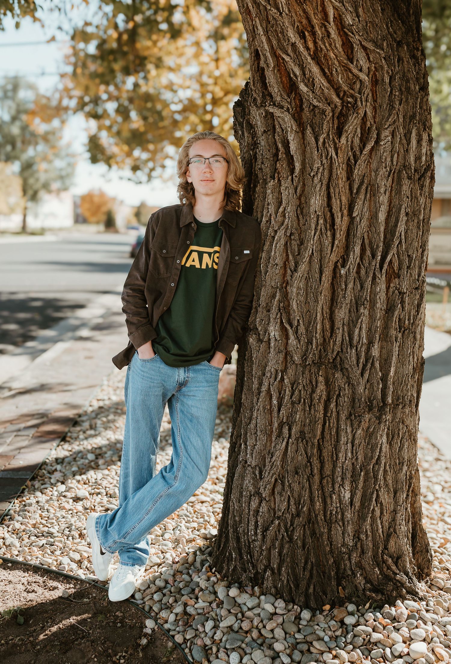 Person in a green sweatshirt and jeans poses casually against a large tree trunk during autumn.