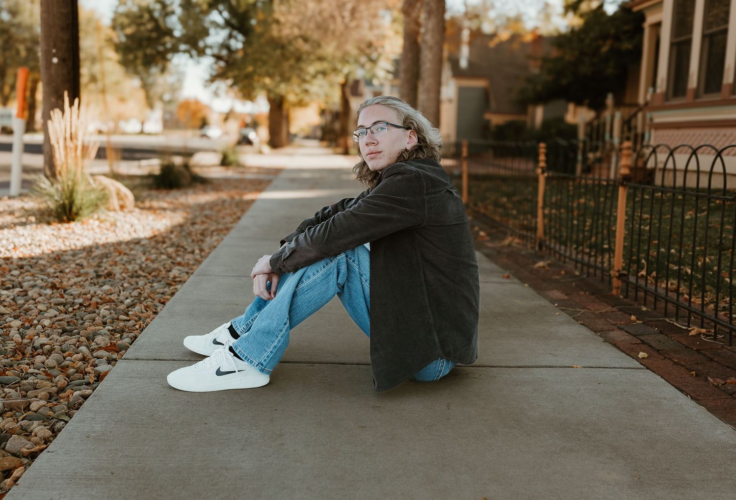 A couple sits together on a sidewalk during autumn with fallen leaves scattered around them.