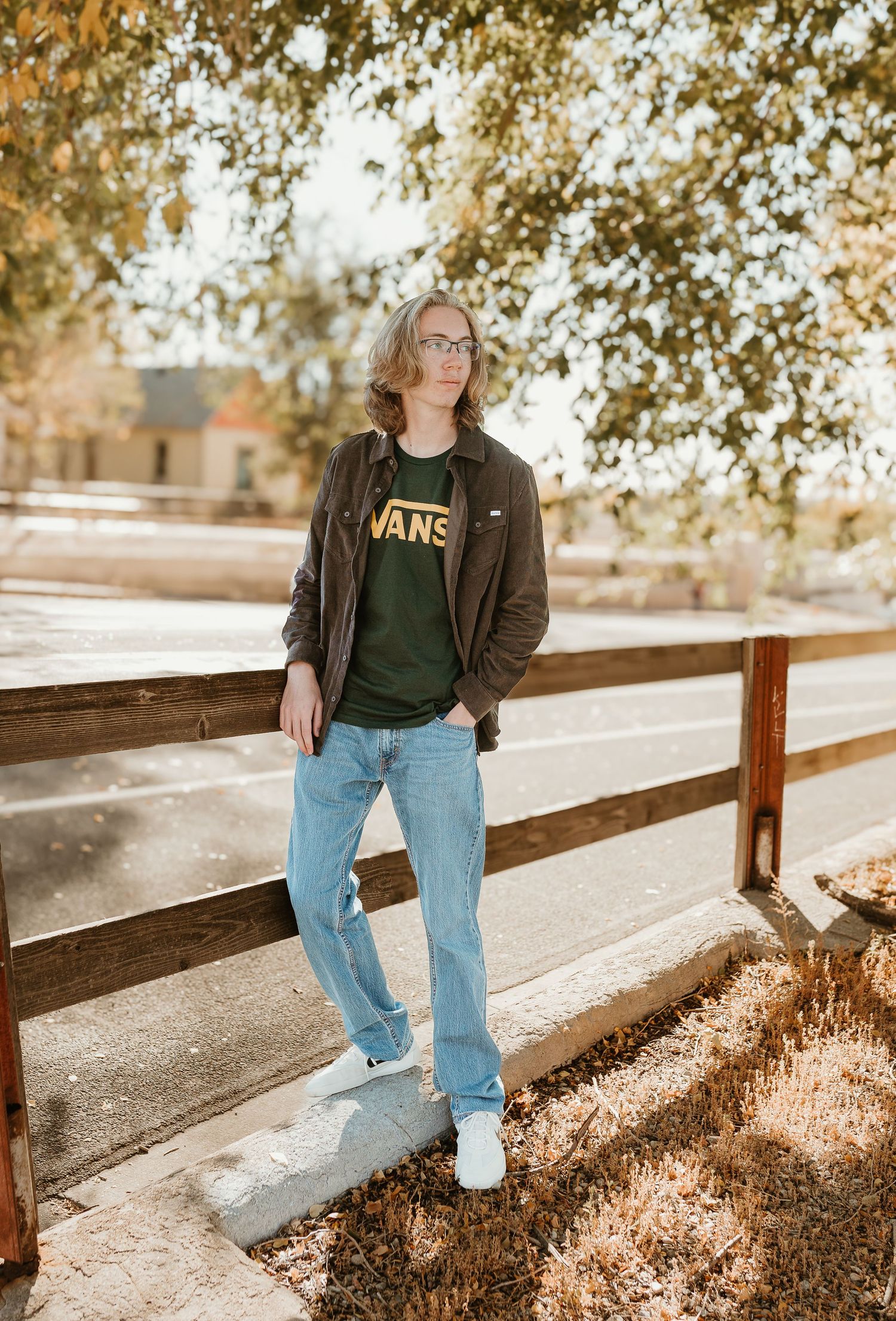 Person in brown jacket and jeans sits on wooden railing in autumn sunlight.