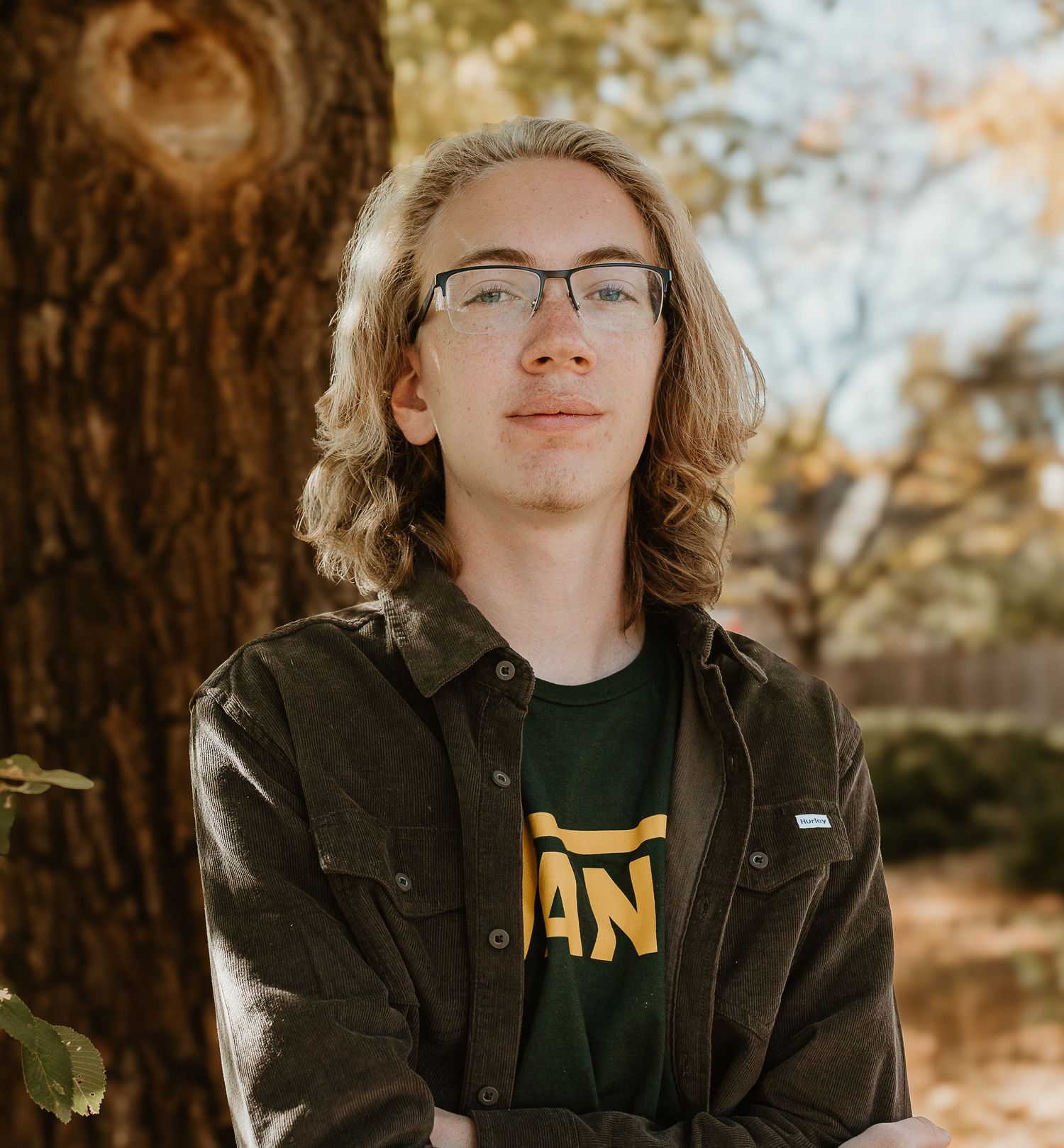 Person in a dark jacket and green shirt poses against an autumn background with warm lighting and trees.