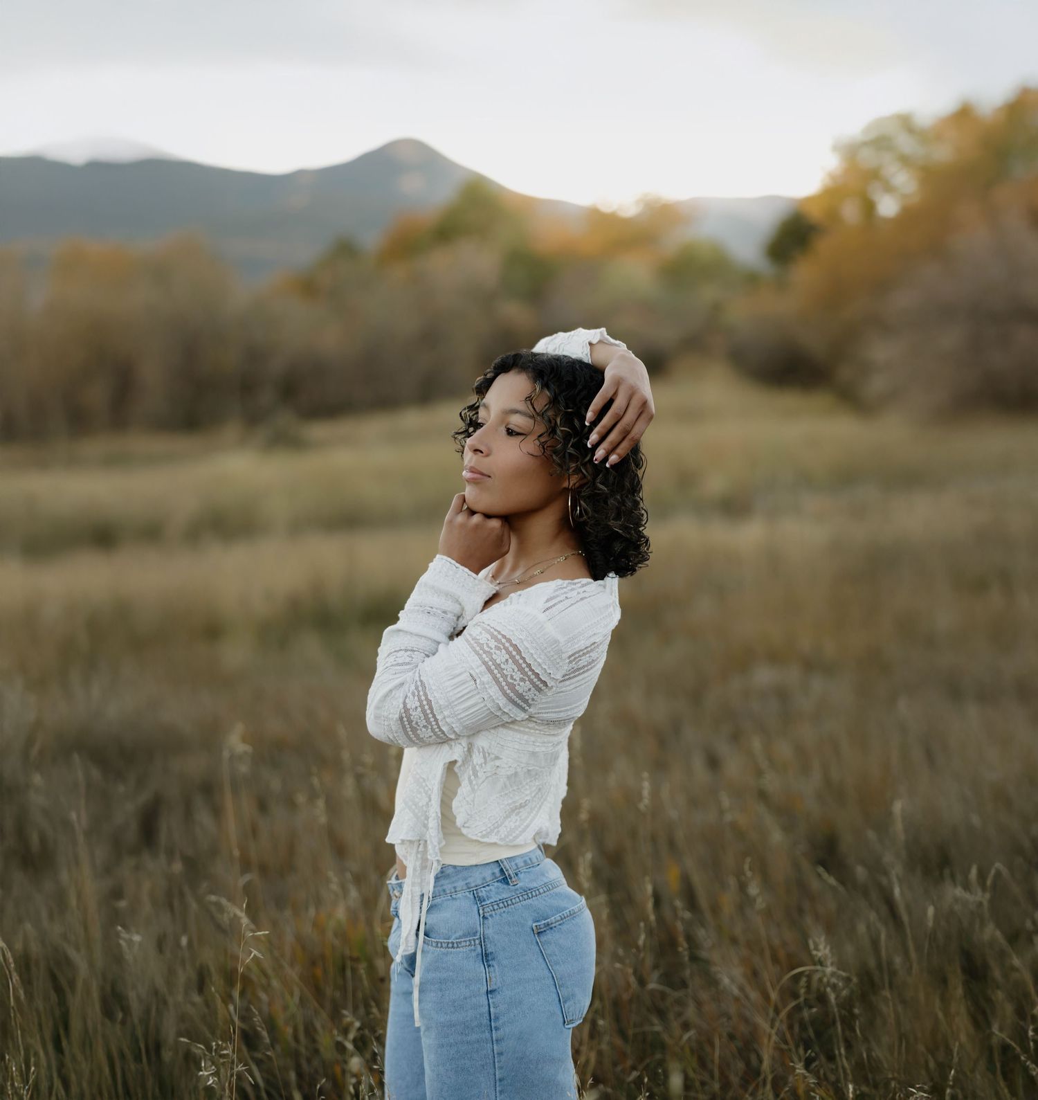 A person in a white sweater and jeans poses thoughtfully in a golden meadow with mountains in the background at sunset.