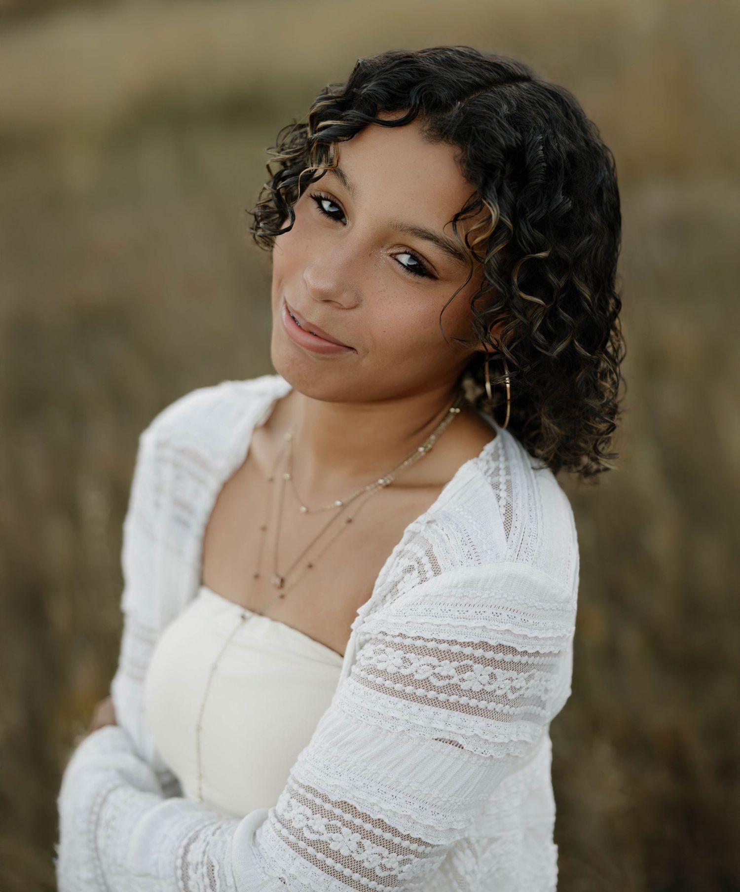 A person in a white lace top with curly hair poses against a blurred outdoor background.