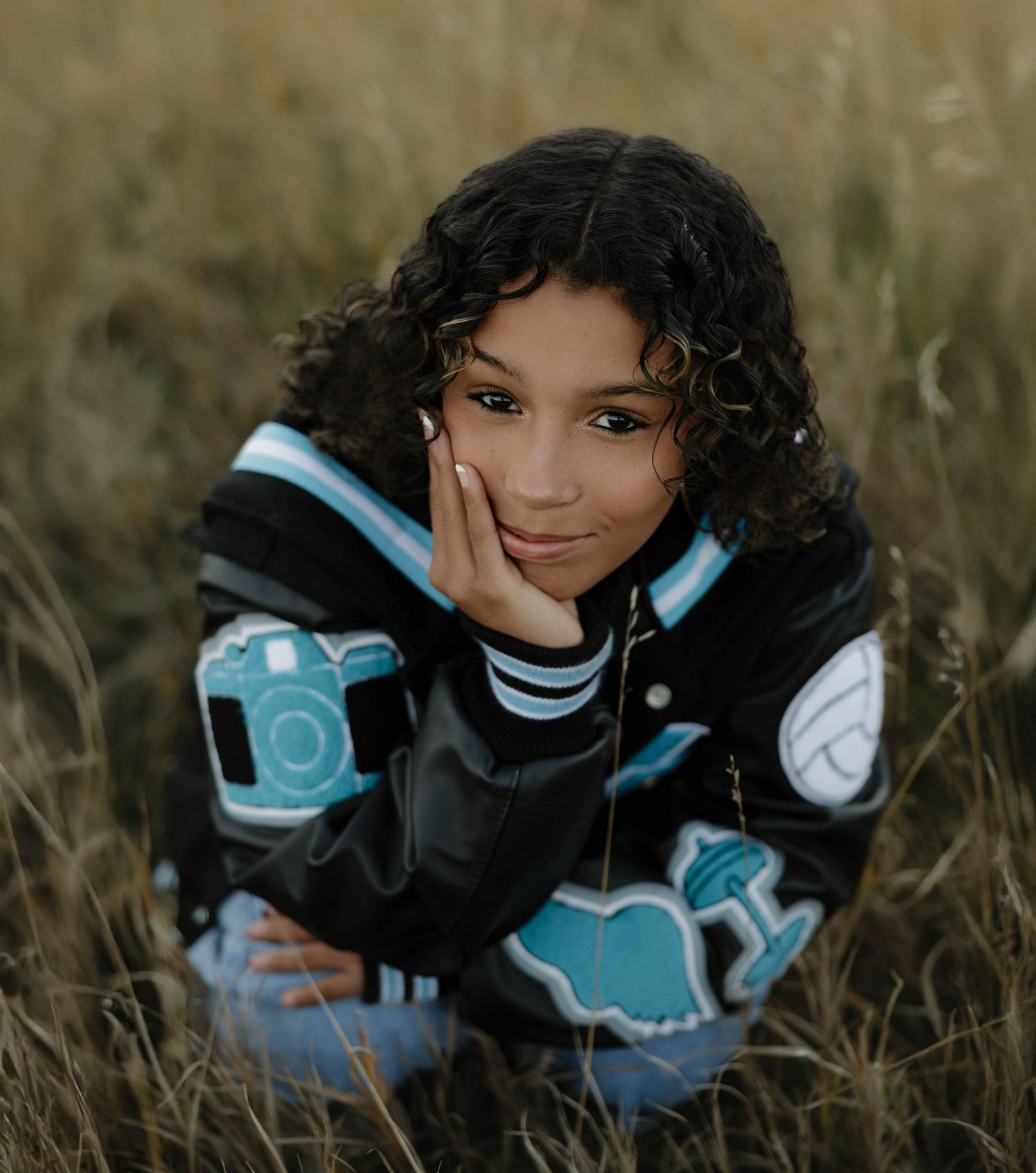 Person wearing black and turquoise jacket sits in tall grass during golden hour outdoor portrait session.