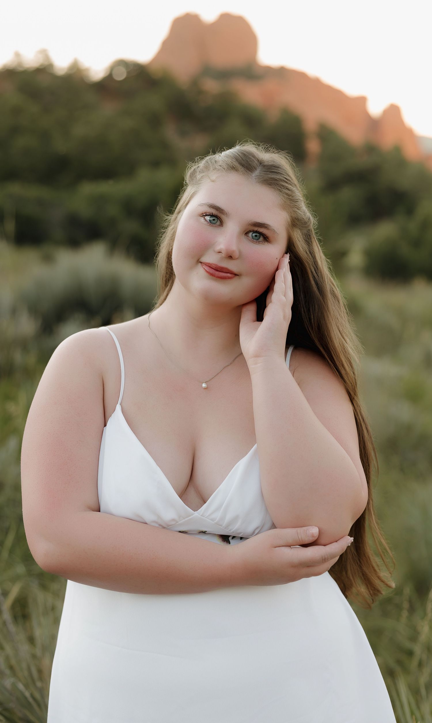 A person in a white top poses against a scenic desert landscape with red rock formations at sunset.