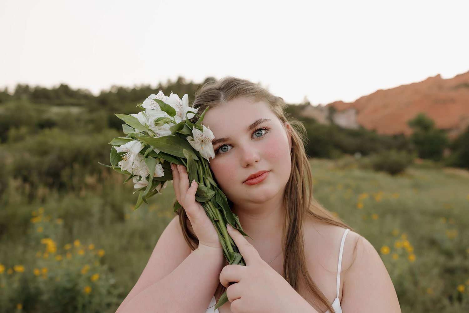 A person poses in a field of wildflowers at sunset holding a white flower near their face.