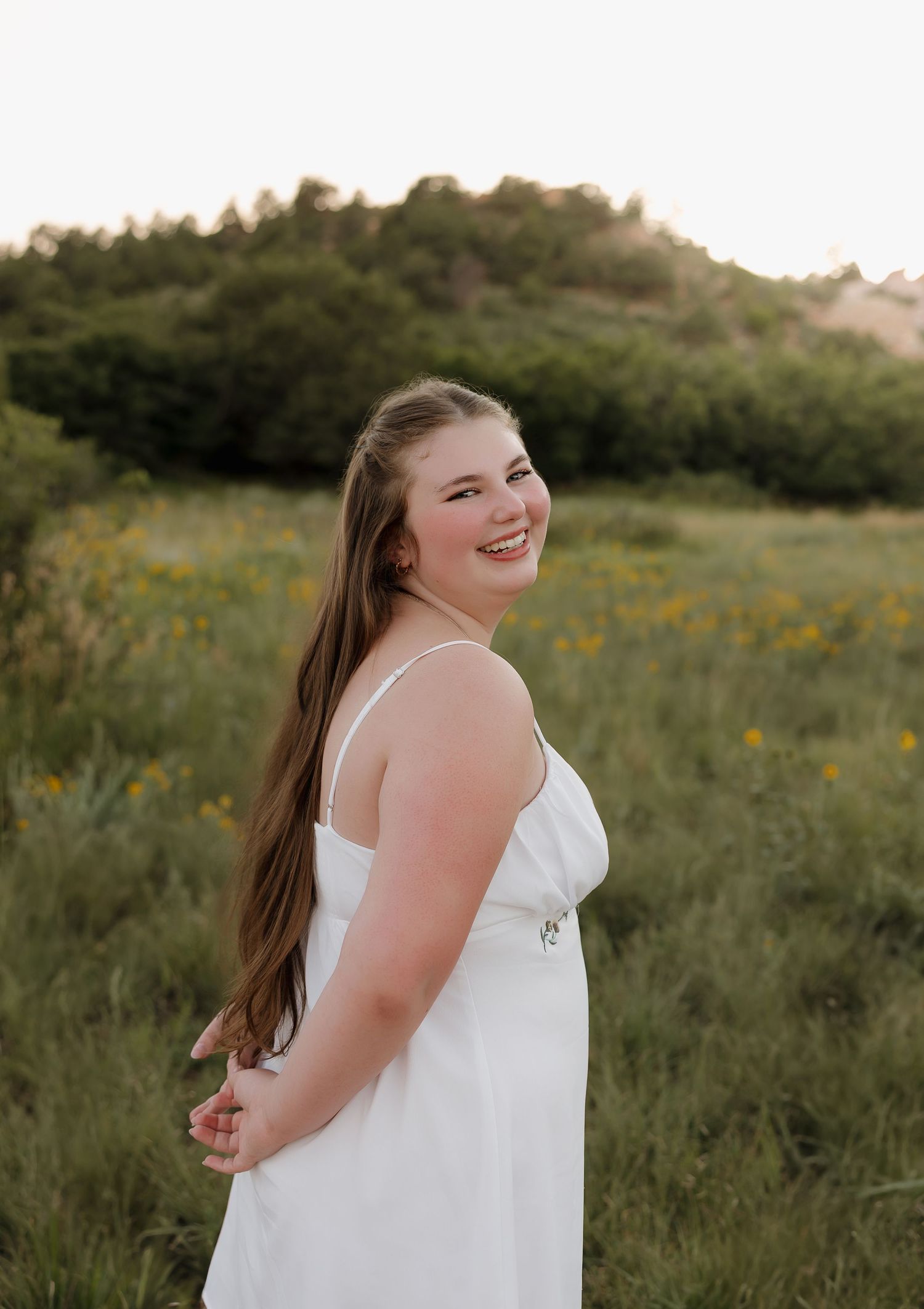 A person in a white sleeveless dress smiles while standing in a grassy field with mountains in the background.