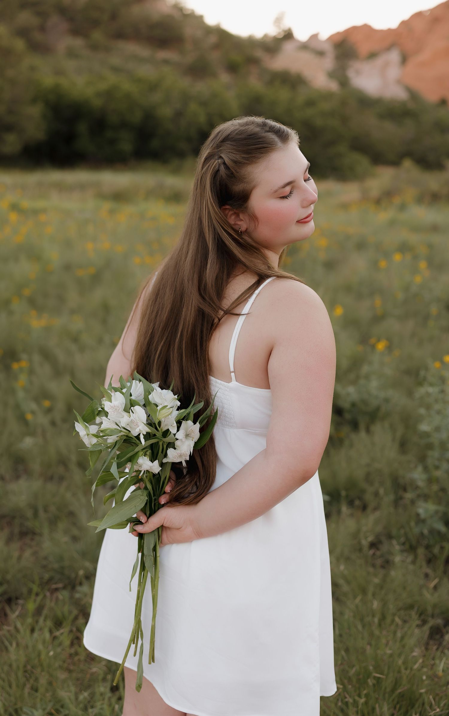 A white floral bouquet is held against a flowing white dress in a sunlit meadow with red rock formations.