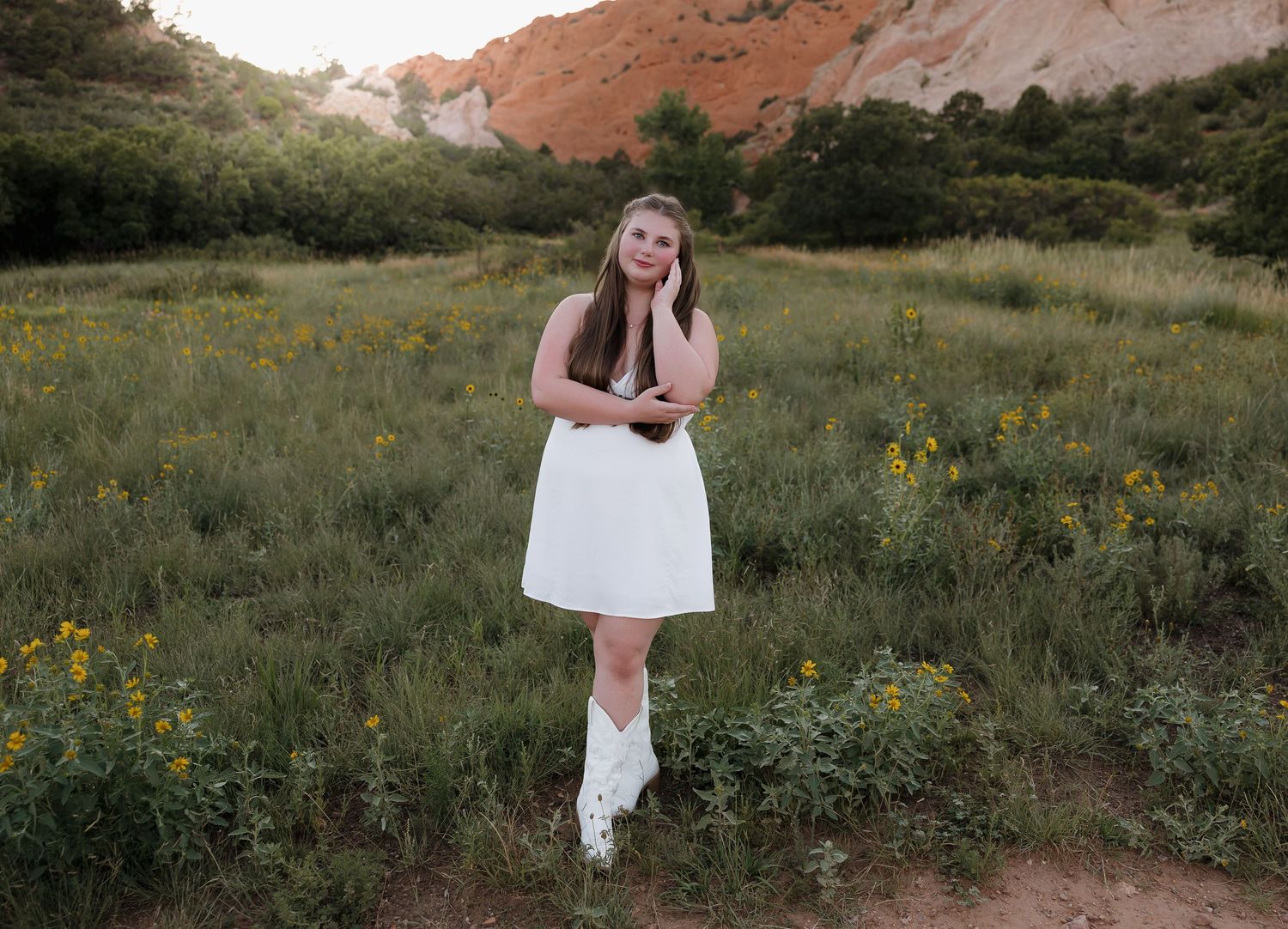 Someone in a white dress and boots stands in a meadow with red rock formations and wildflowers in the background.