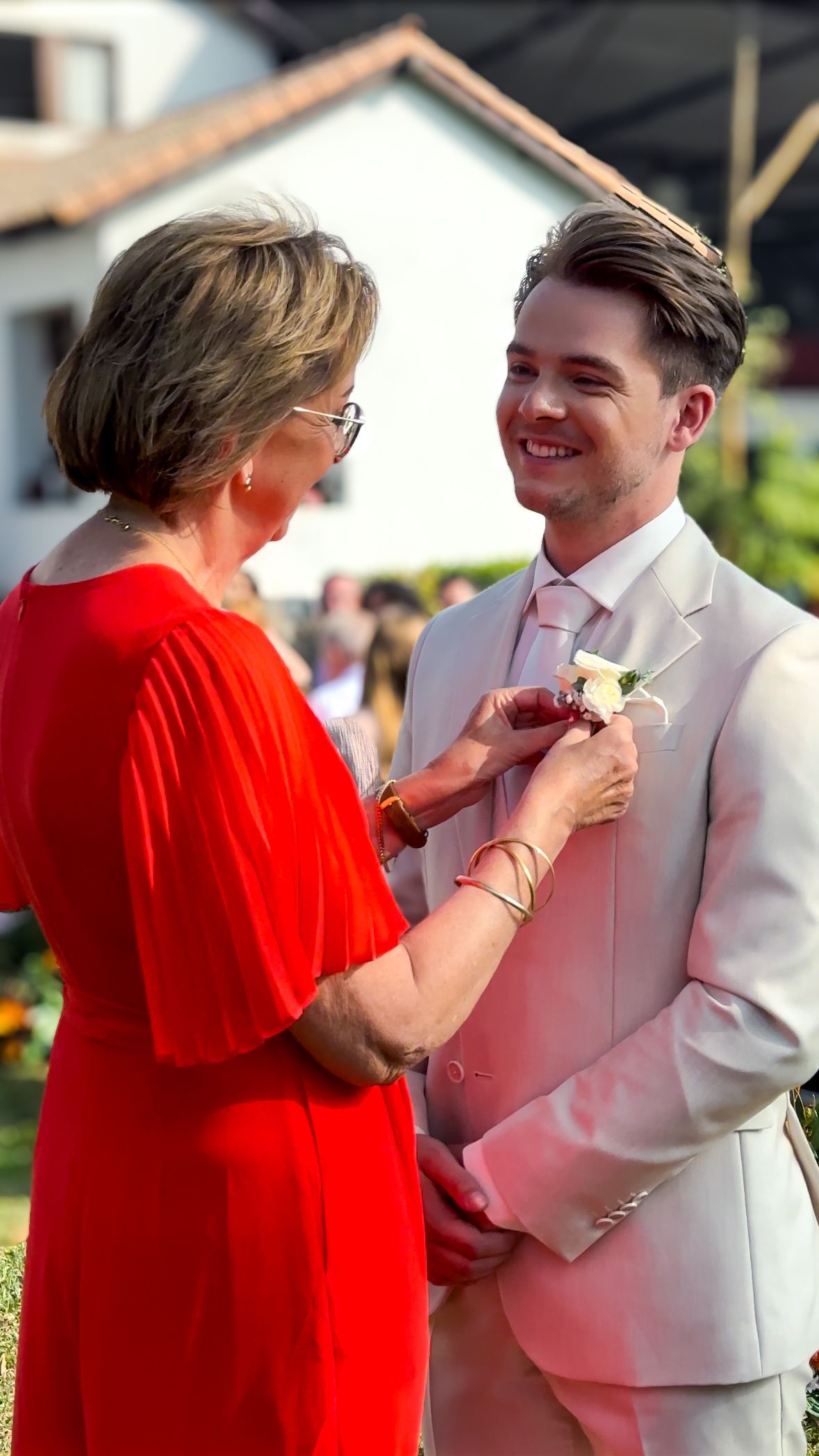Person in red dress pinning boutonniere on someone wearing white formal attire outdoors.