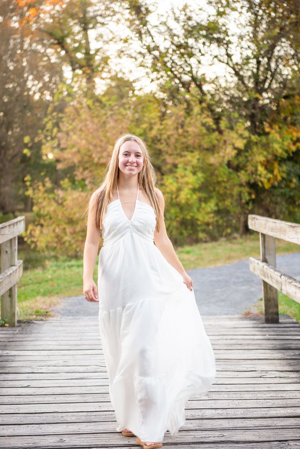 A person in a flowing white dress twirls on a wooden boardwalk surrounded by autumn foliage in soft sunlight.