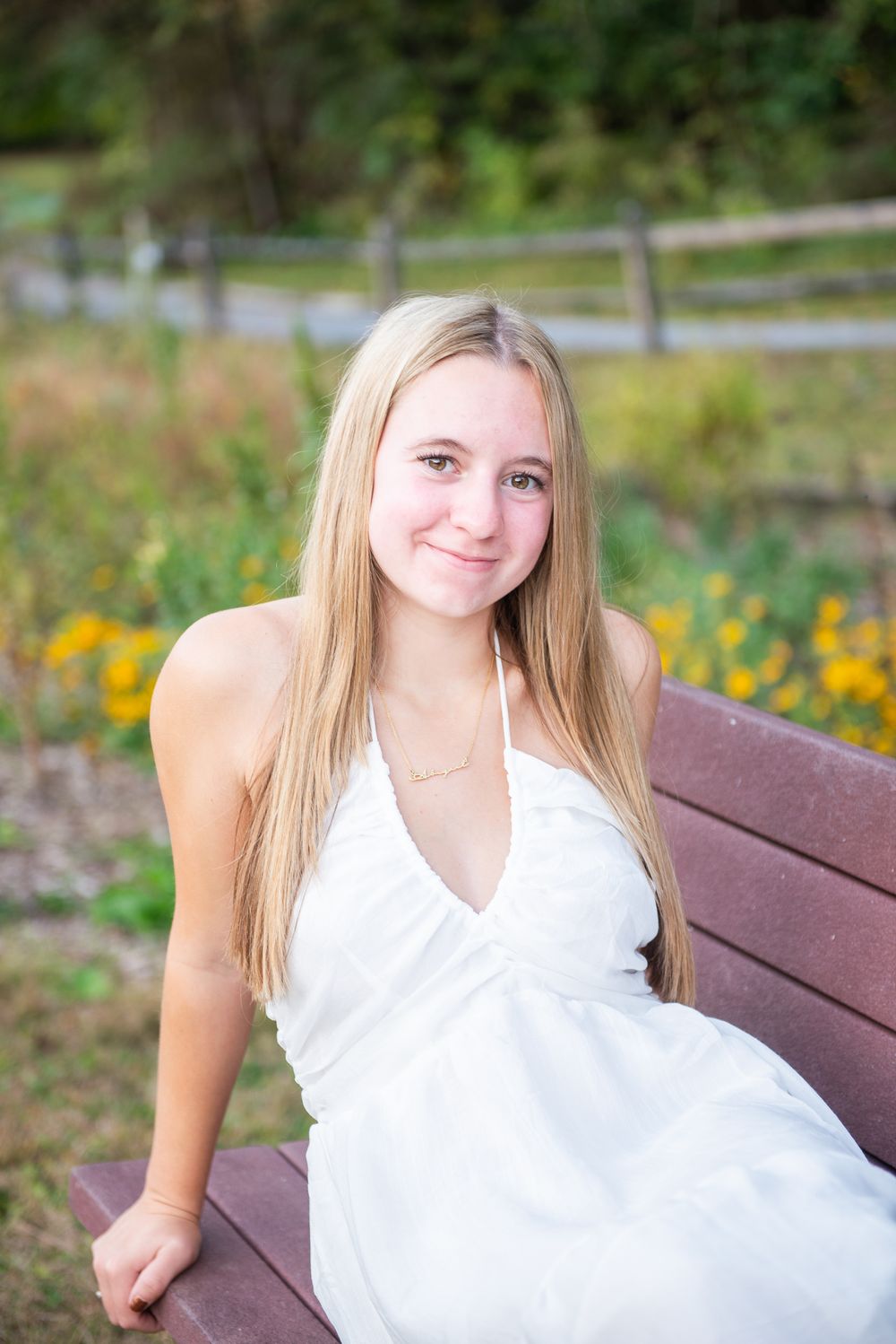 A person in a white summer dress sits on a wooden bench in a garden setting with yellow flowers in the background.