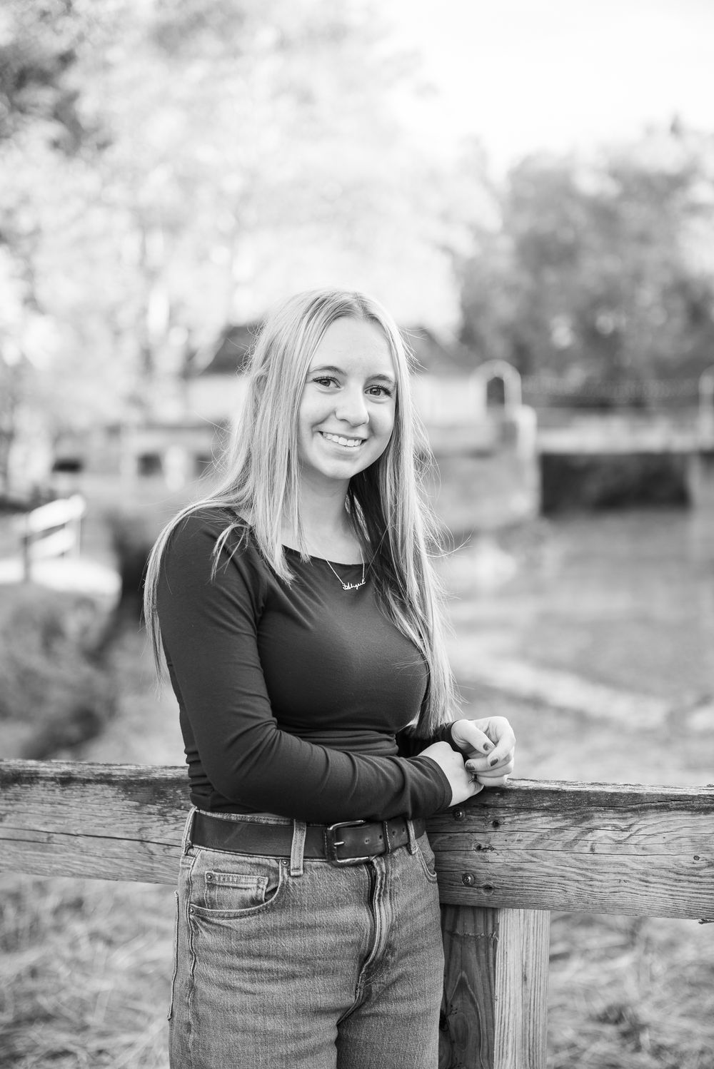 A person in a black long-sleeve top and jeans leans against a wooden fence in a black and white outdoor portrait.