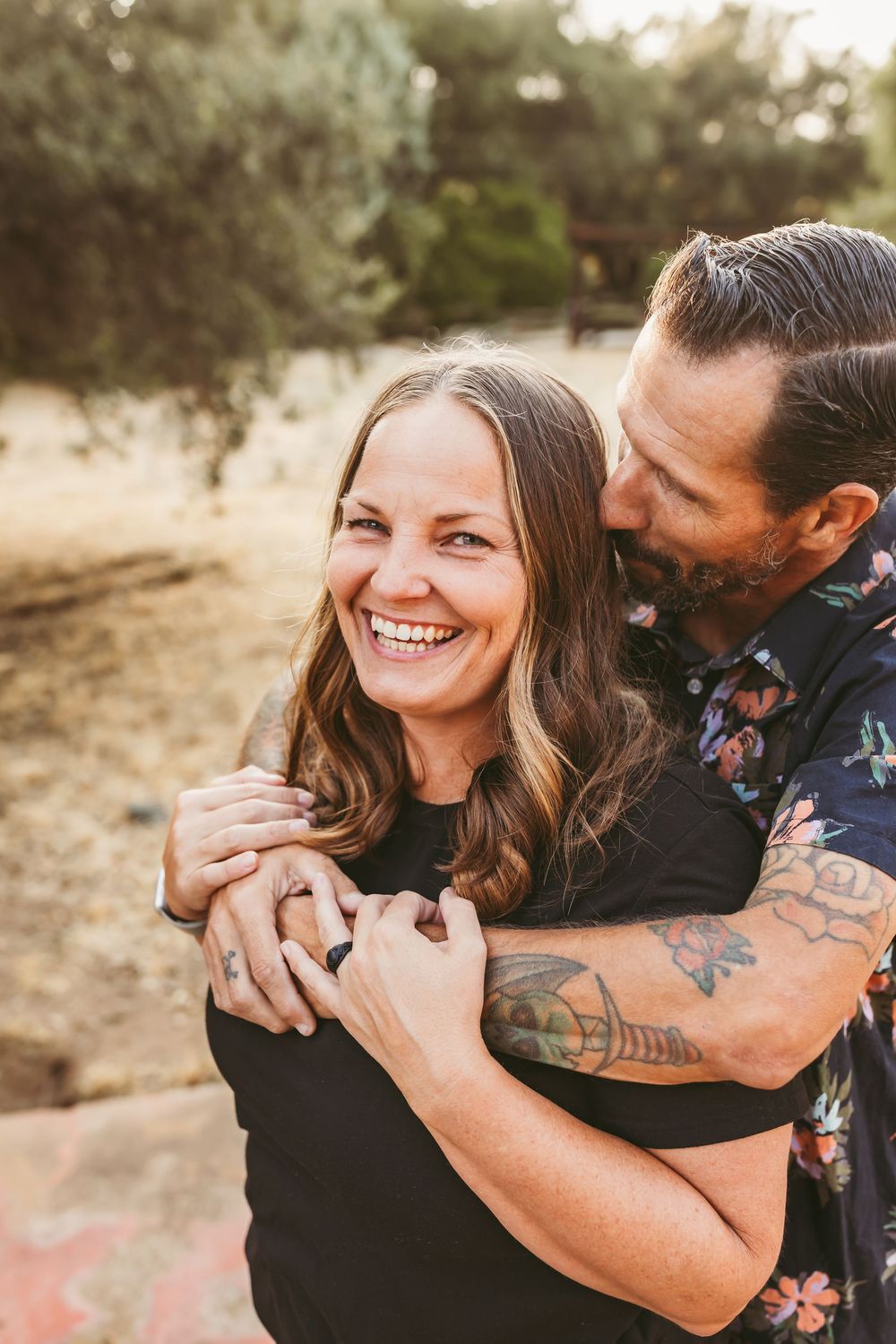 A couple embraces and laughs together during a golden hour photoshoot in a natural outdoor setting.
