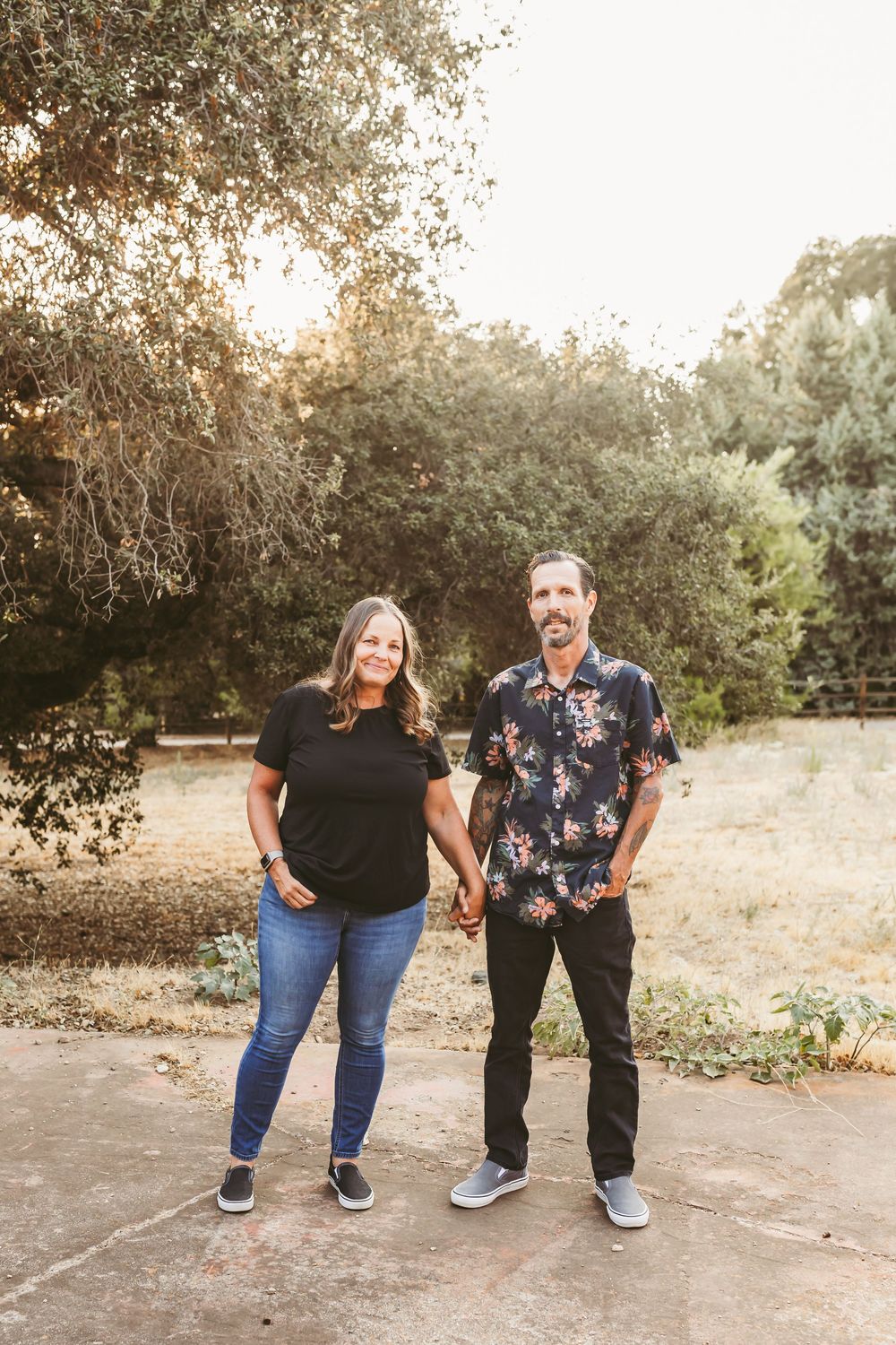 A couple stands together outdoors on a dirt path surrounded by trees during golden hour.