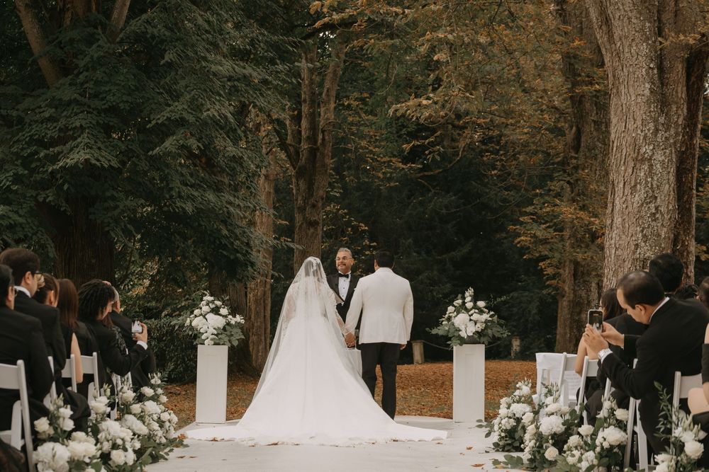 Outdoor wedding ceremony setup in a wooded area with white chairs and floral arrangements.