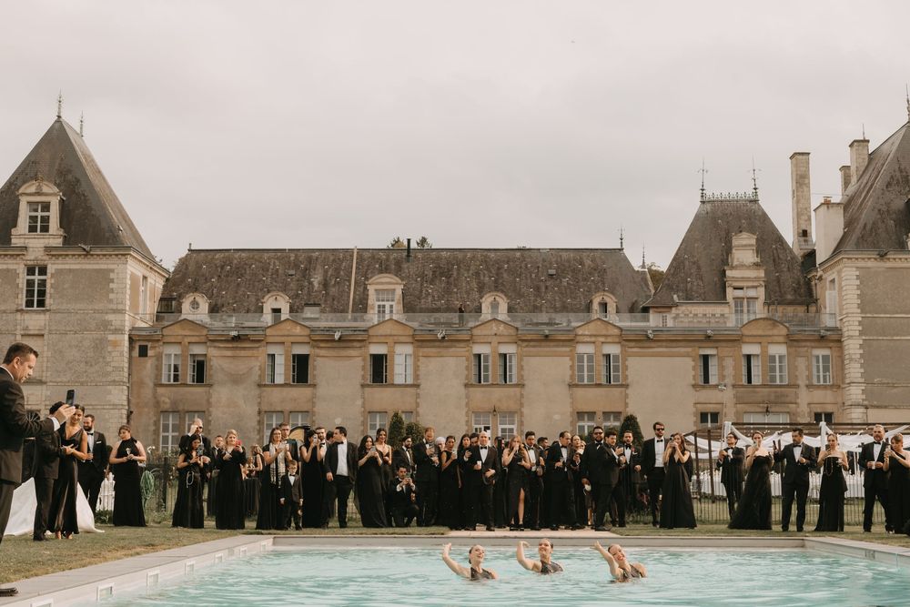 Elegant wedding party gathered in front of a grand French chateau on a cloudy day.