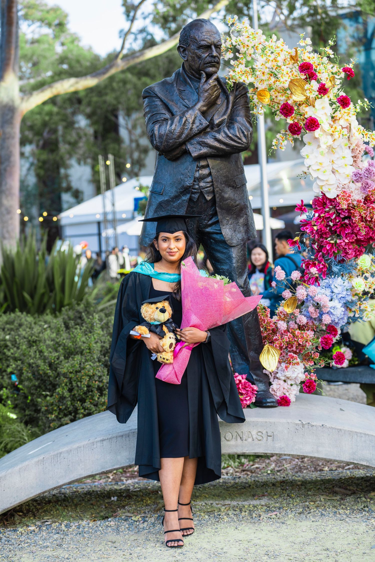 A graduate in black cap and gown holds a vibrant bouquet of pink and red flowers next to a campus statue.