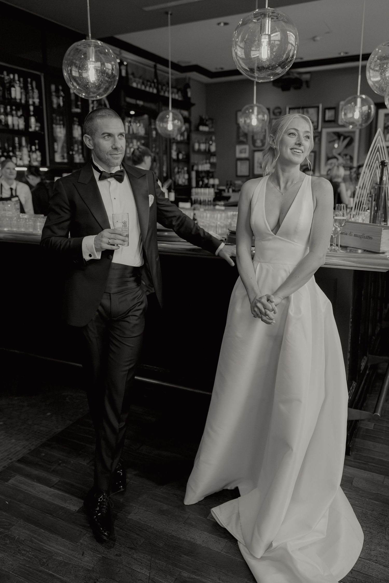 Elegant bride and groom in formal attire pose together at a bar in black and white wedding portrait.
