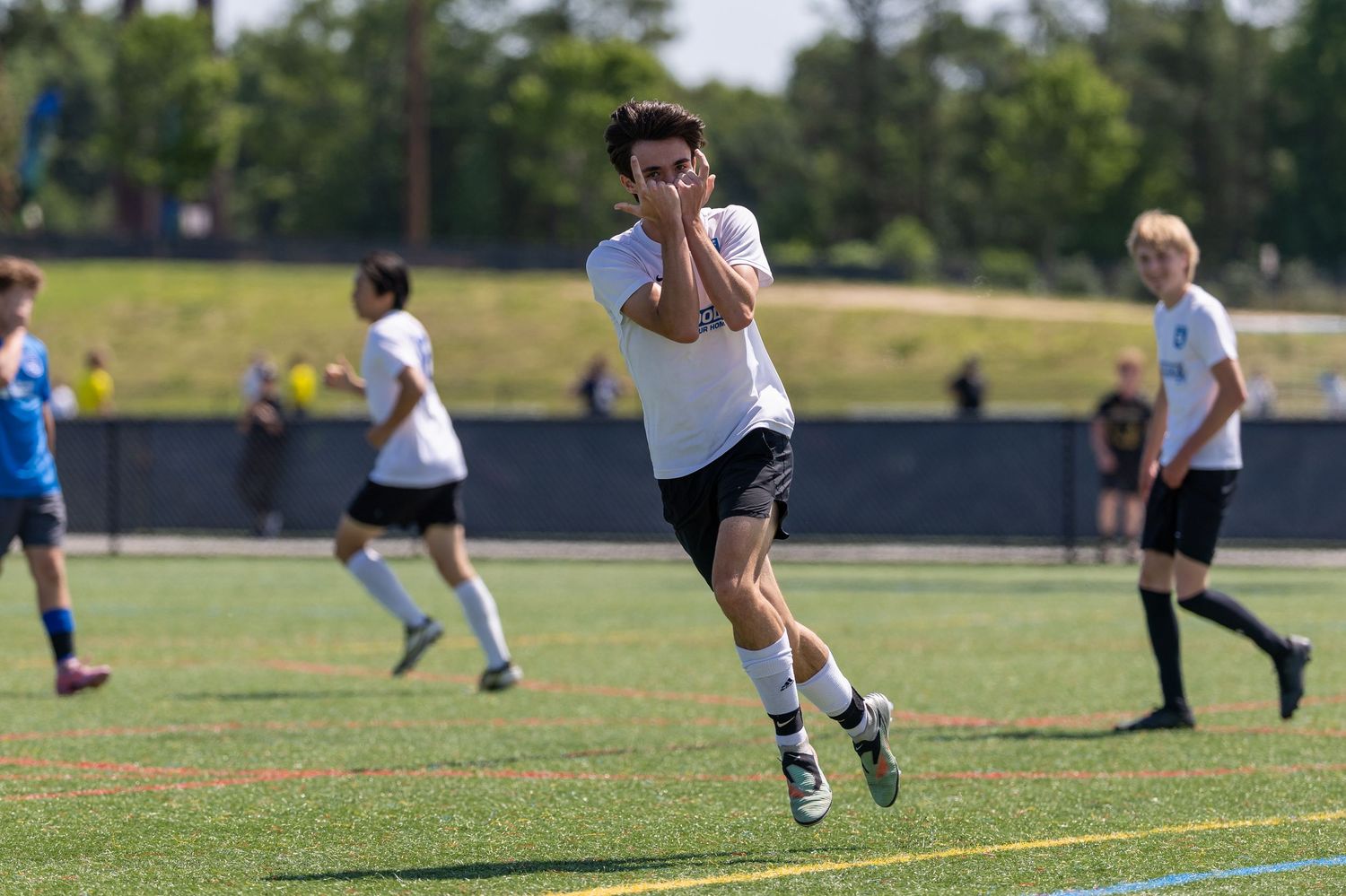 Soccer players compete for the ball on a sunny grass field with goal posts visible in background.