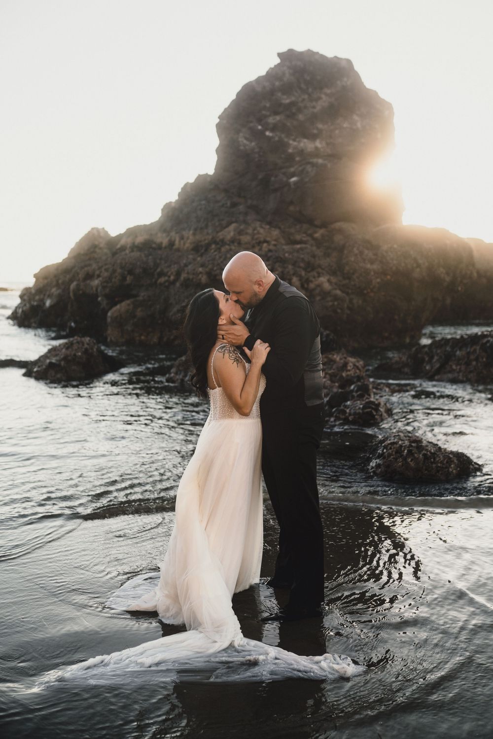 A romantic couple embraces during sunset at a rocky beach coastline with waves crashing around them.