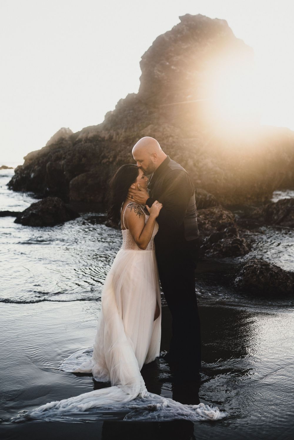 A romantic couple embraces on a rocky beach at sunset with waves and cliffs creating a dramatic silhouette.