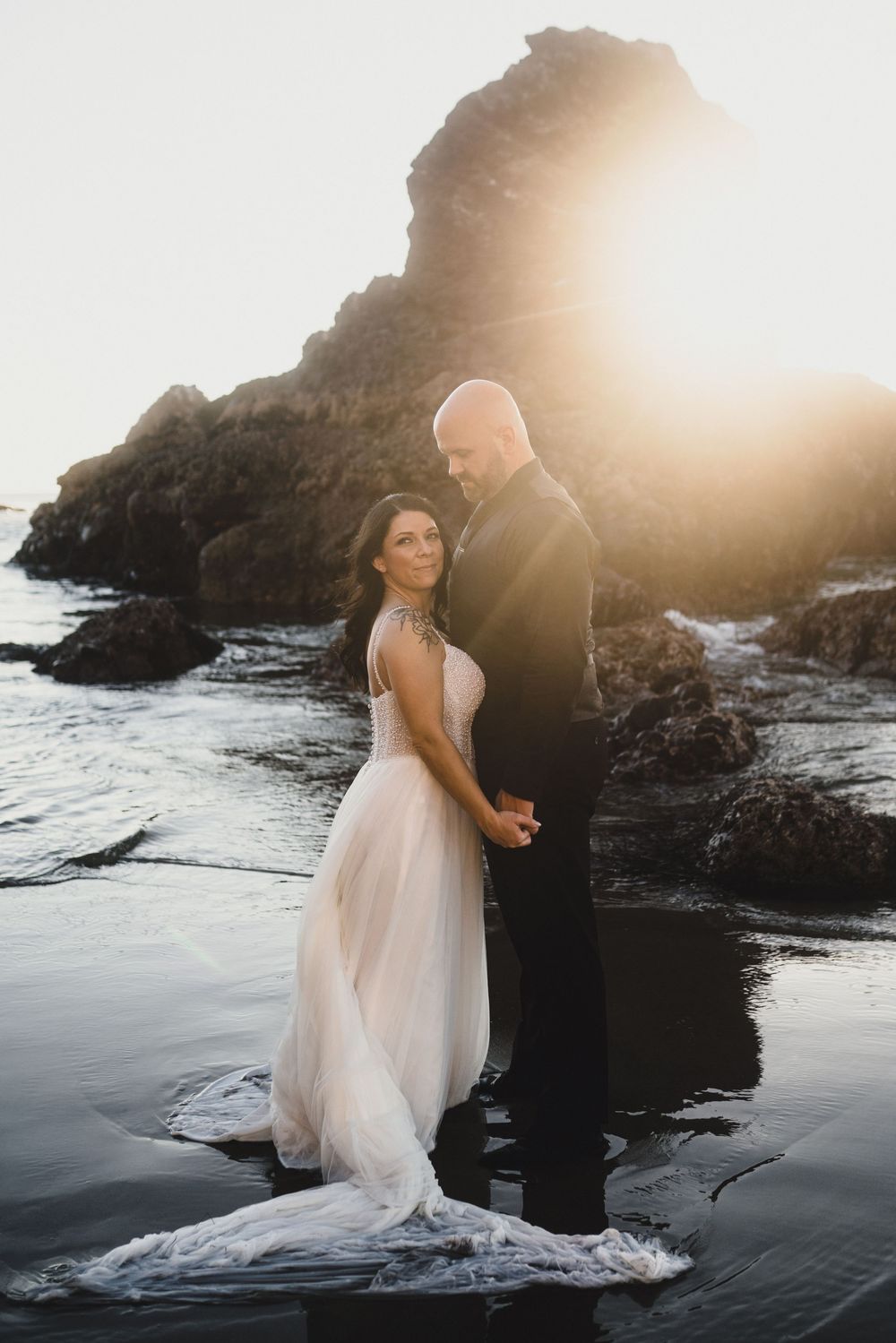 A couple embraces on a rocky beach at sunset with dramatic ocean waves and cliffs in the background.