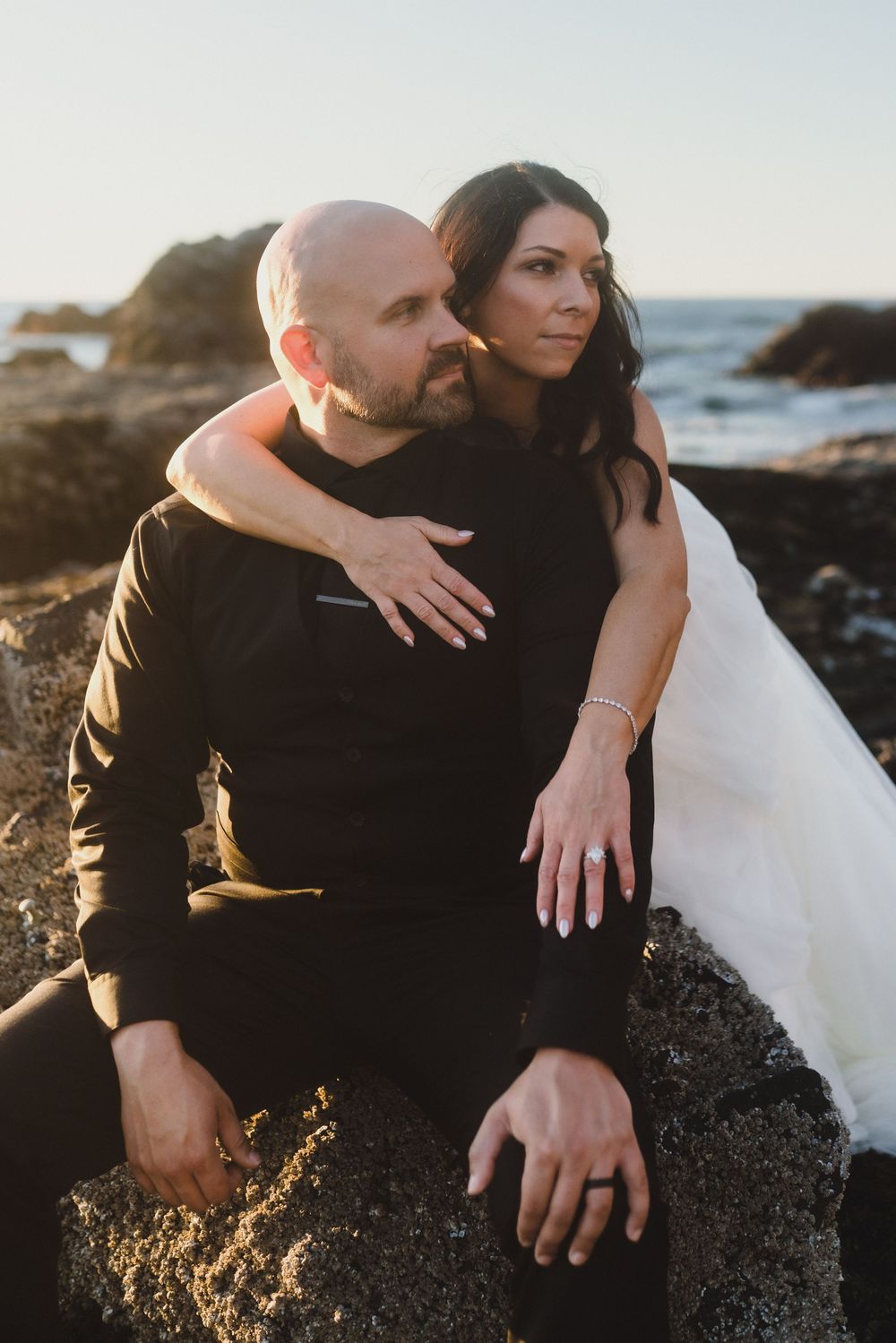 A couple embraces on rocky coastal cliffs during golden hour sunset with ocean waves crashing in the background.