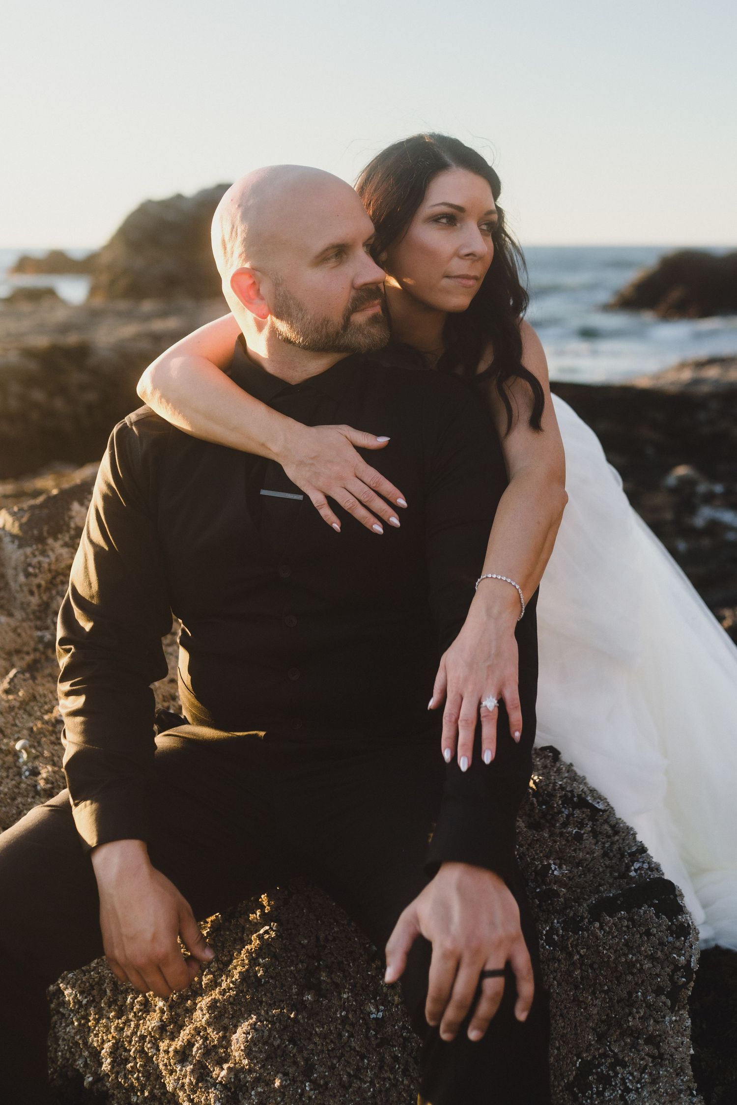 A couple embraces on rocky coastal cliffs during golden hour sunset with ocean waves crashing in the background.