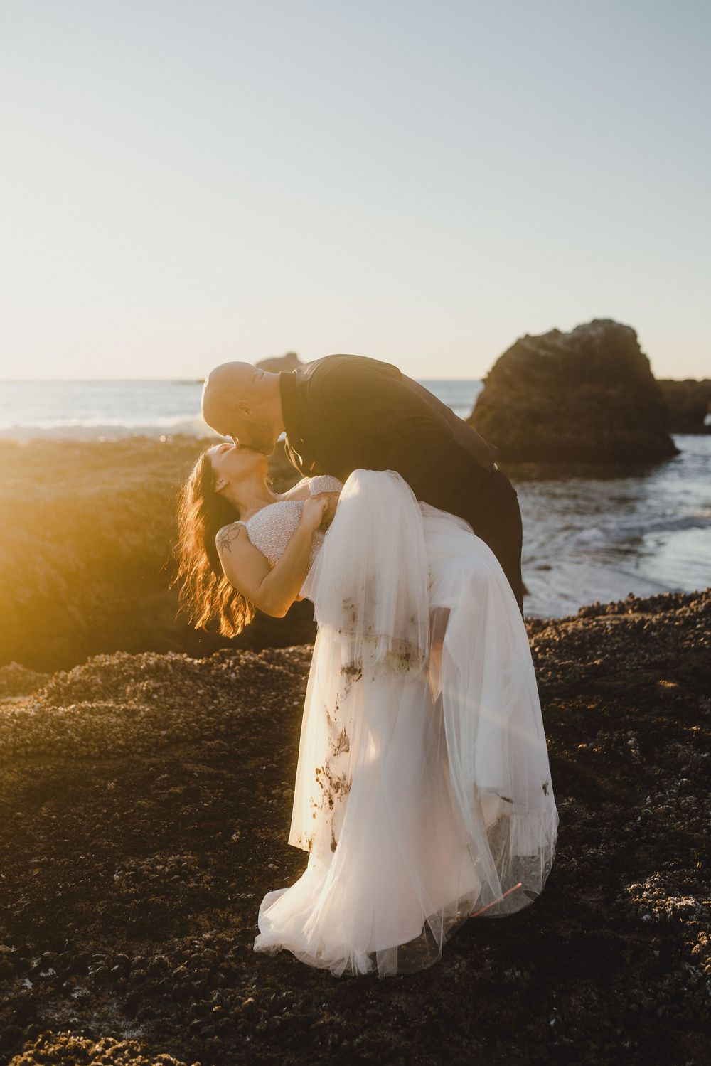 A romantic seaside photoshoot with a flowing white wedding dress against dramatic coastal rocks at sunset.