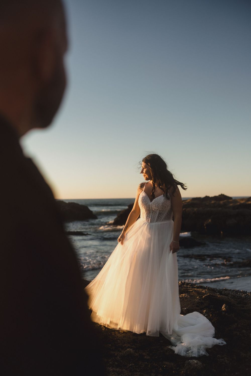 A bride in a flowing white tulle gown stands on rocky coastal cliffs at sunset while a silhouette watches from afar.