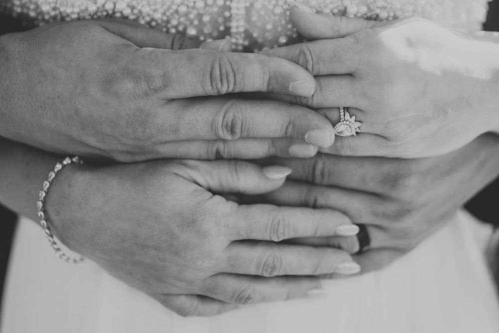 Close-up black and white photo of hands with wedding rings resting together in a romantic pose.