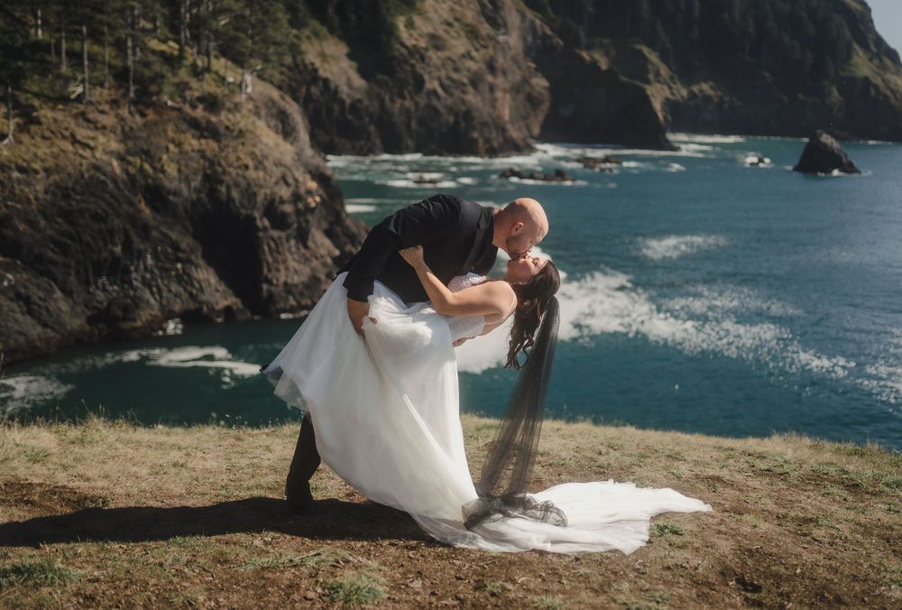 A couple in wedding attire sits together on a scenic coastal cliff overlooking dramatic ocean views and rocky shoreline.