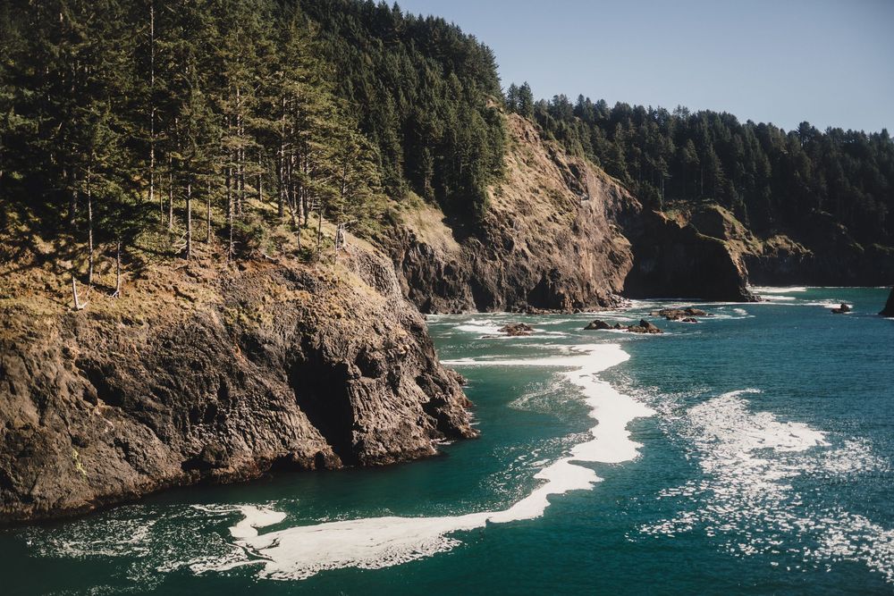 Dramatic coastal cliffs meet turquoise waters along the rugged Oregon coastline with evergreen forest above.