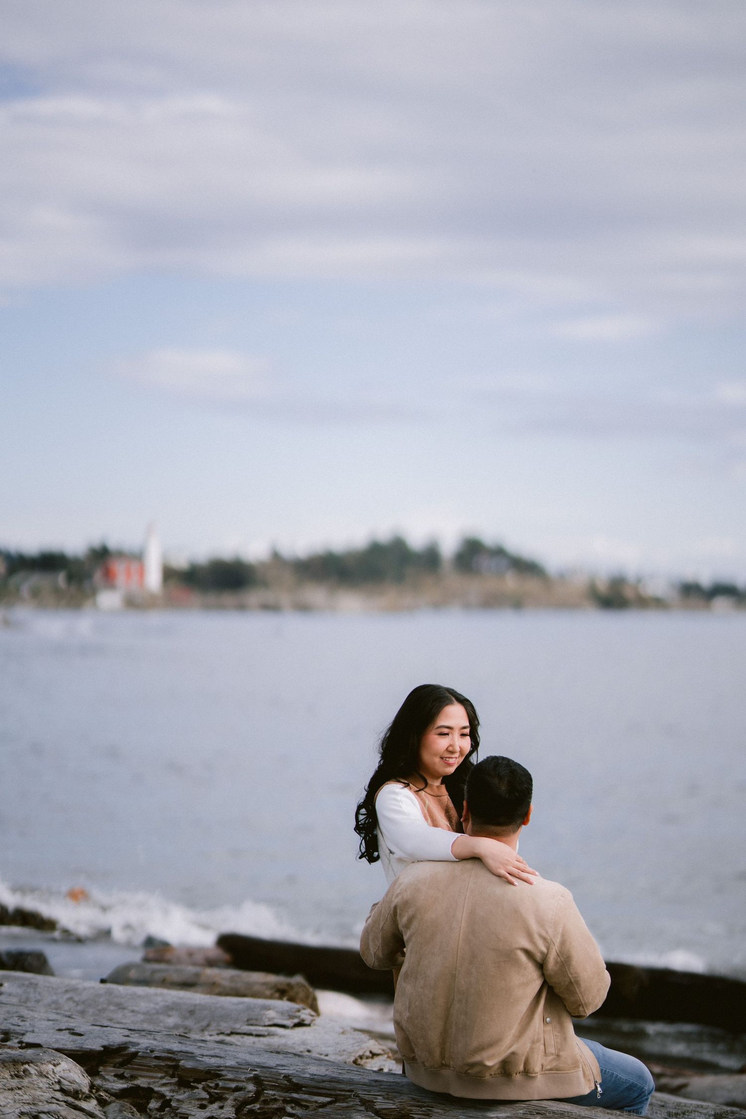 A couple sitting together on coastal rocks overlooking a waterfront with buildings in the distance.