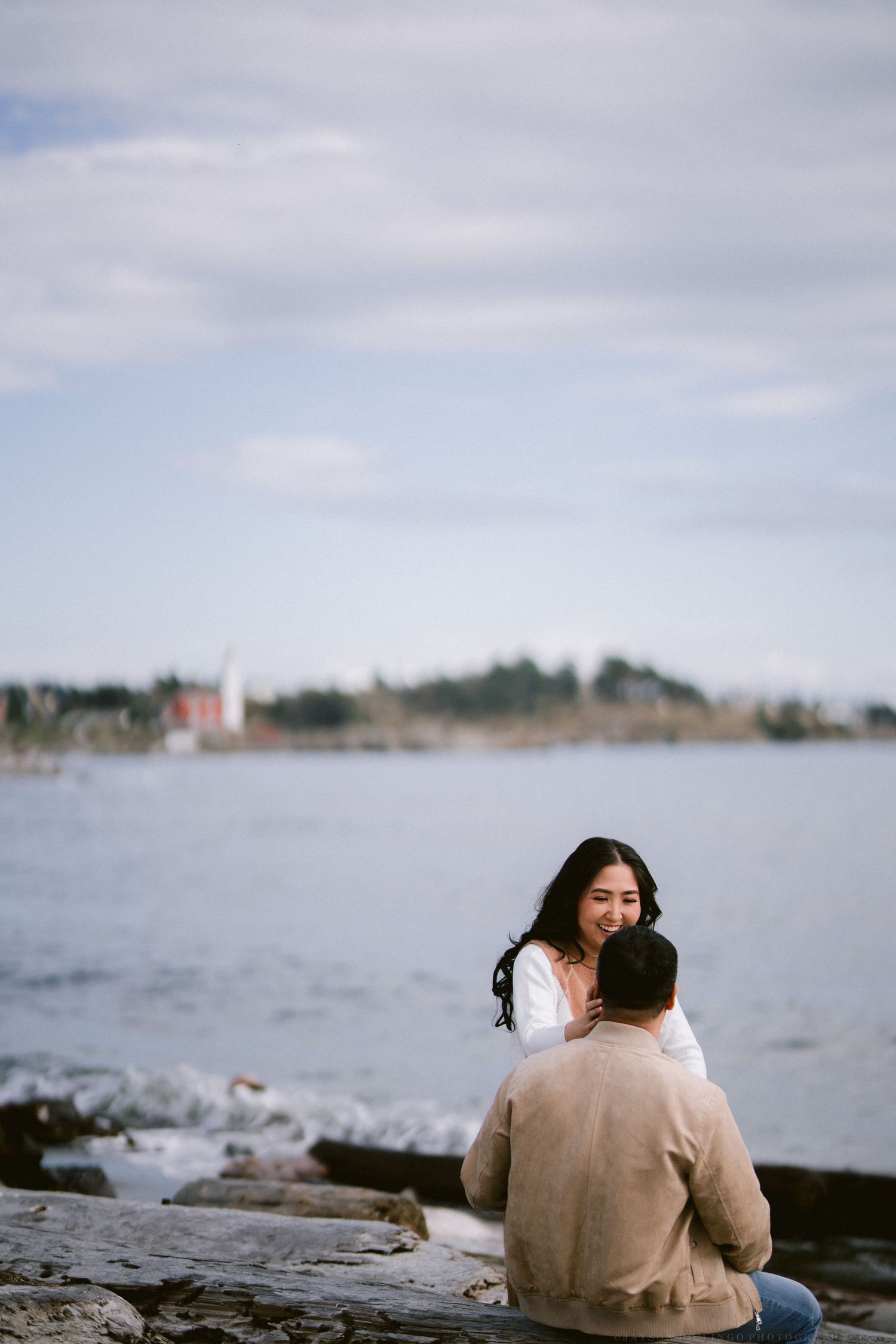 Couple sits together on rocky shoreline overlooking calm ocean waters on overcast day.