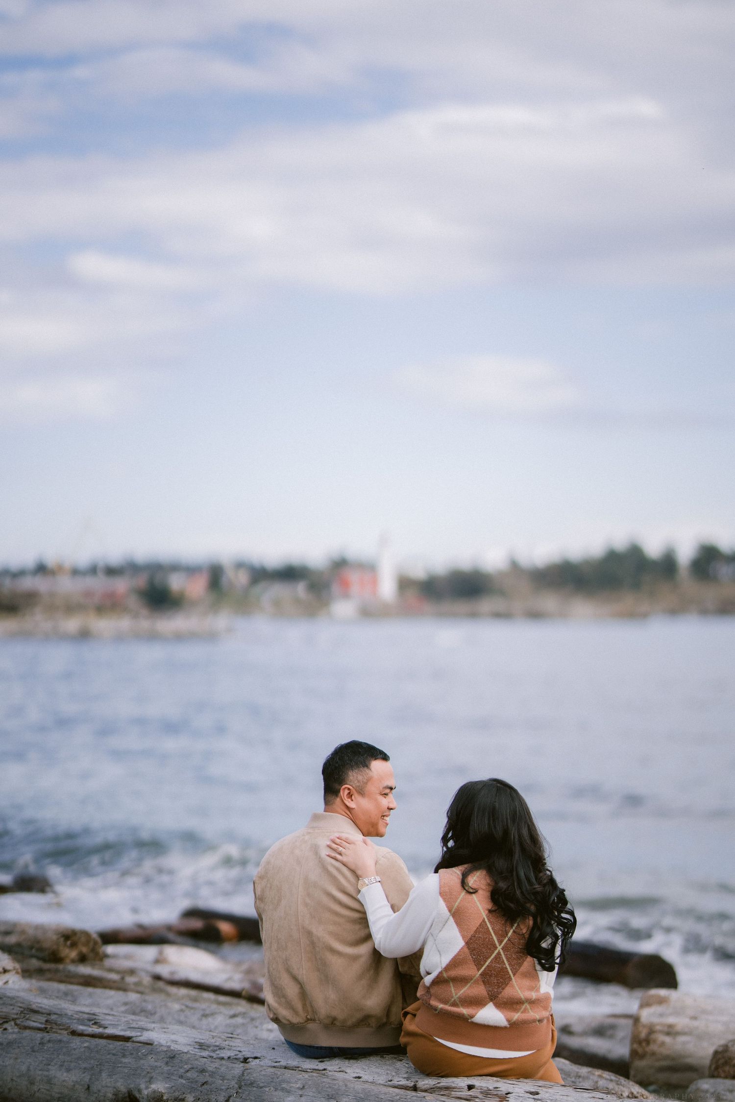A couple sits together on coastal rocks overlooking a scenic waterfront on a cloudy day.