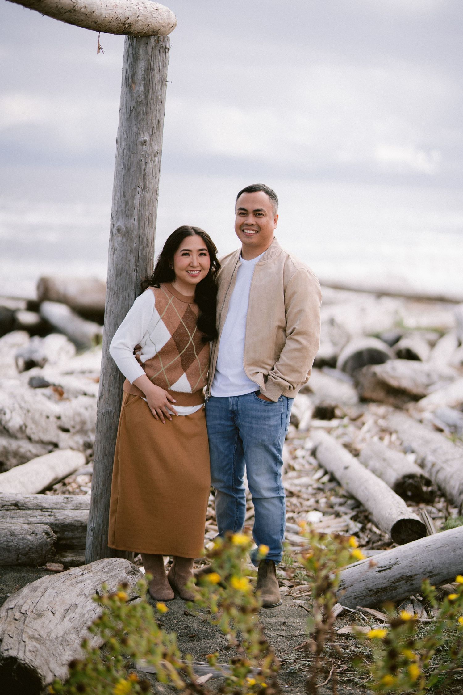 A couple poses together on driftwood by the shore wearing casual earth-toned clothing.