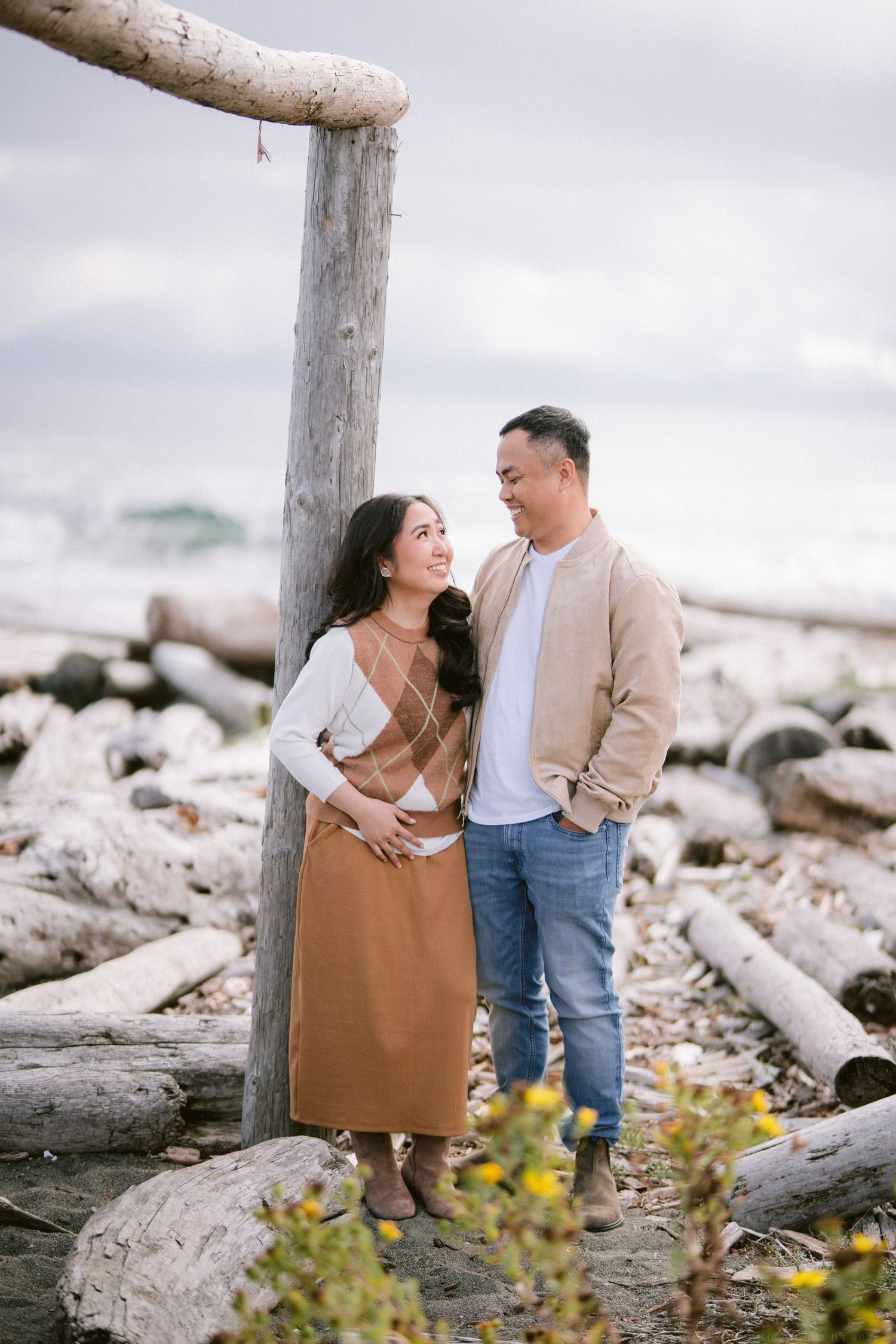 A couple shares a romantic moment among driftwood logs on a rocky beach with cloudy skies in the background.